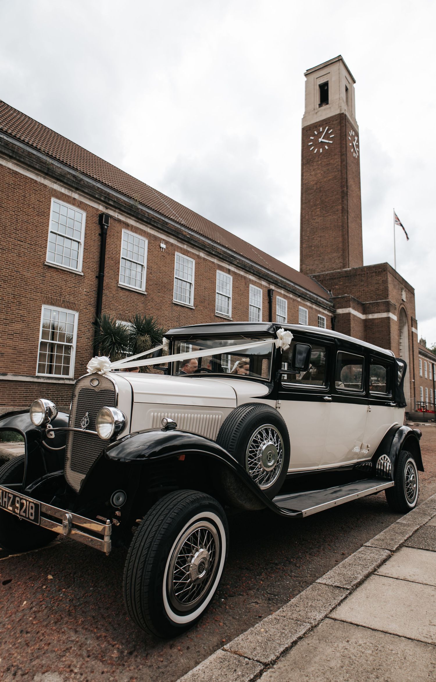 Vintage white and black wedding car decorated with ribbons parked outside a brick building with clock tower.