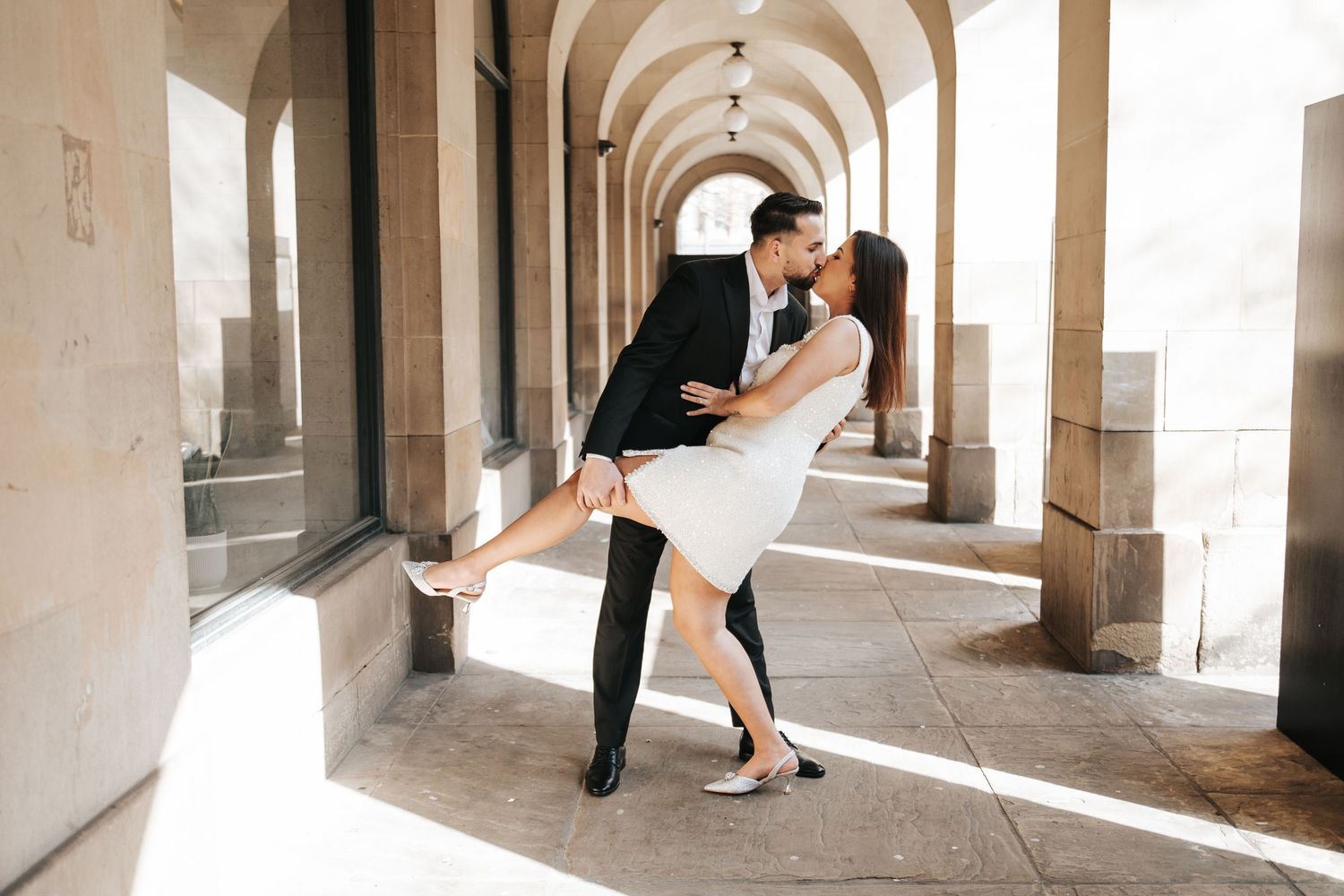 Couple sharing a romantic dip kiss under arched colonnade, woman in white dress, man in black suit.