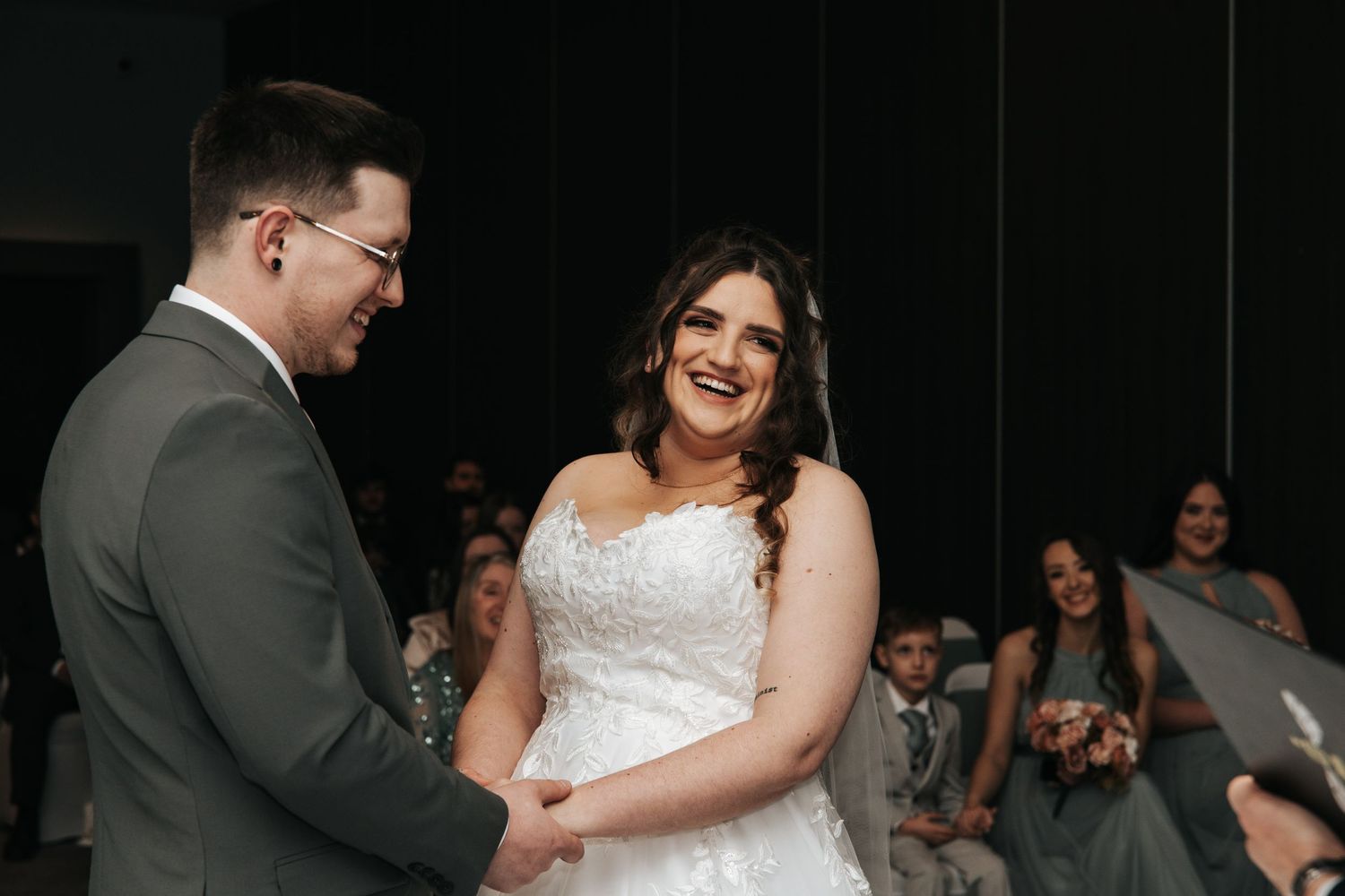 Bride in white lace gown laughs joyfully while holding hands with groom in gray suit during wedding ceremony.