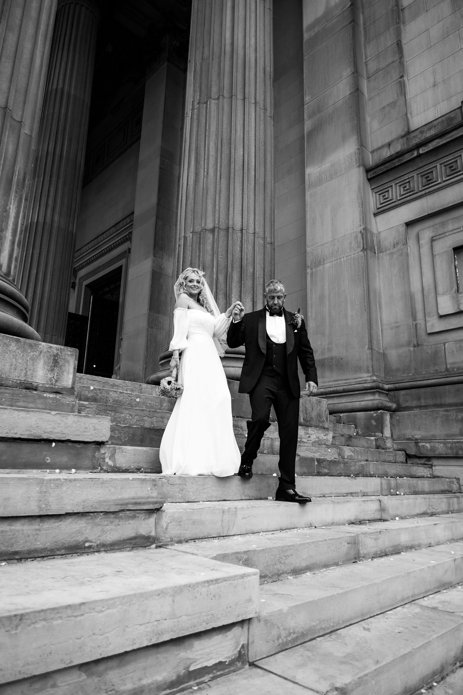 Black and white photo of a bride and groom holding hands on grand stone steps with tall columns behind them.
