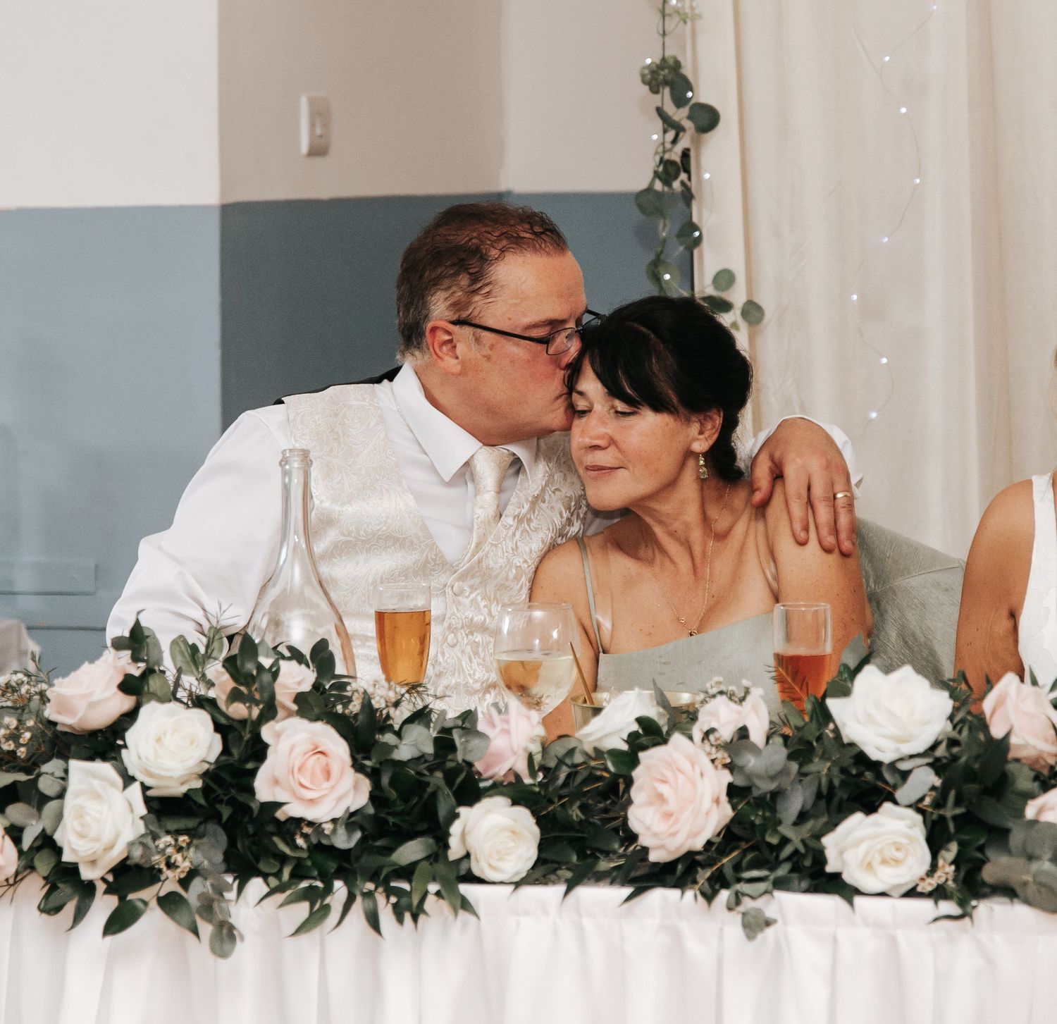 Bride and groom share a tender kiss at wedding reception table adorned with white and pink floral arrangements.