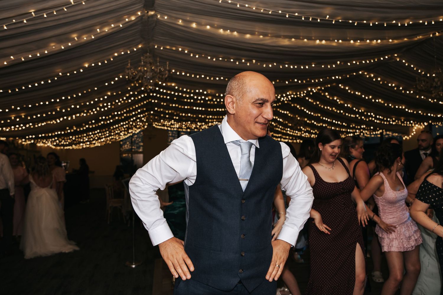 Bald man in navy vest dancing confidently at elegant wedding reception.
