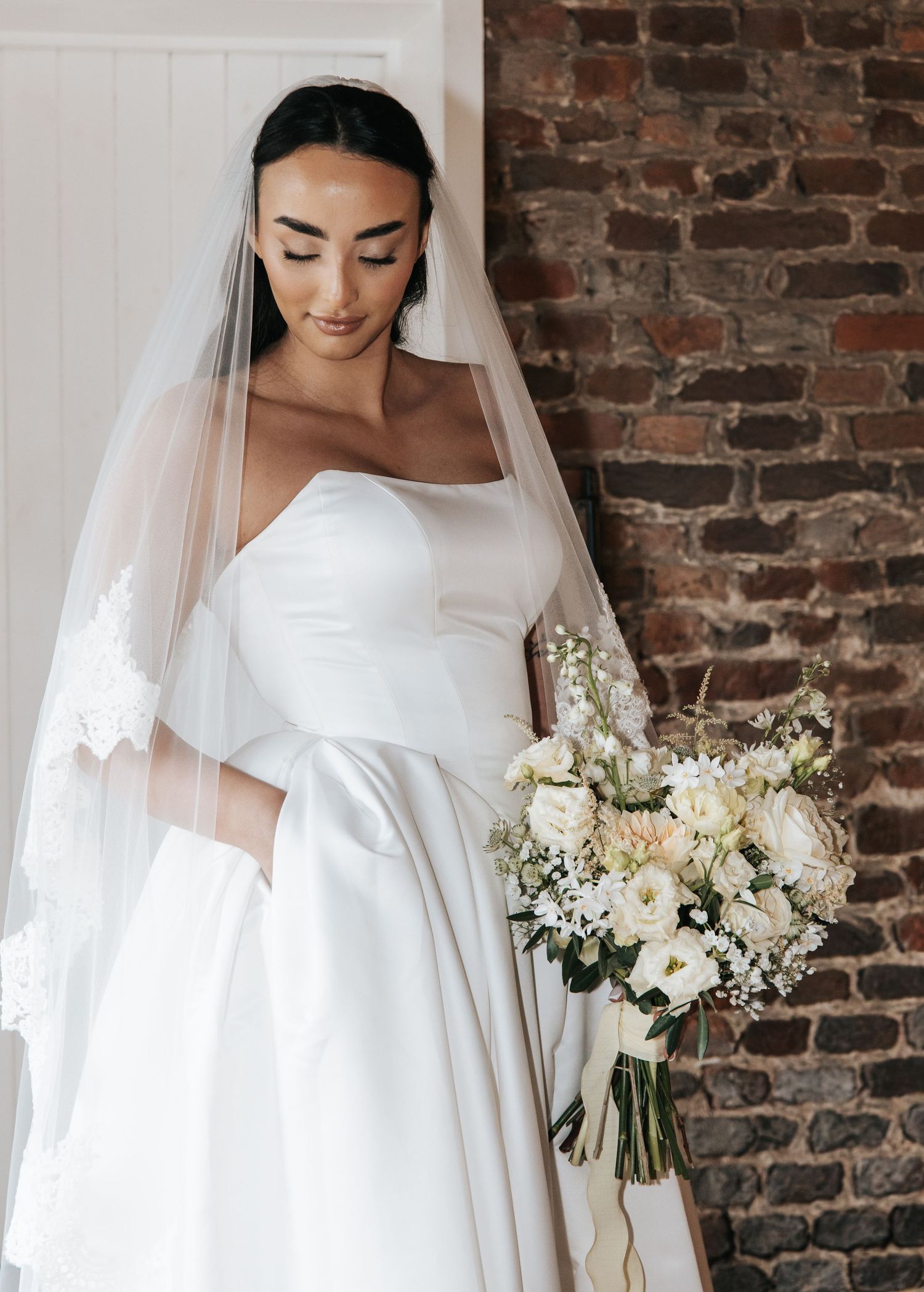 Elegant bride in classic white strapless ballgown and veil poses with white floral bouquet against exposed brick wall.
