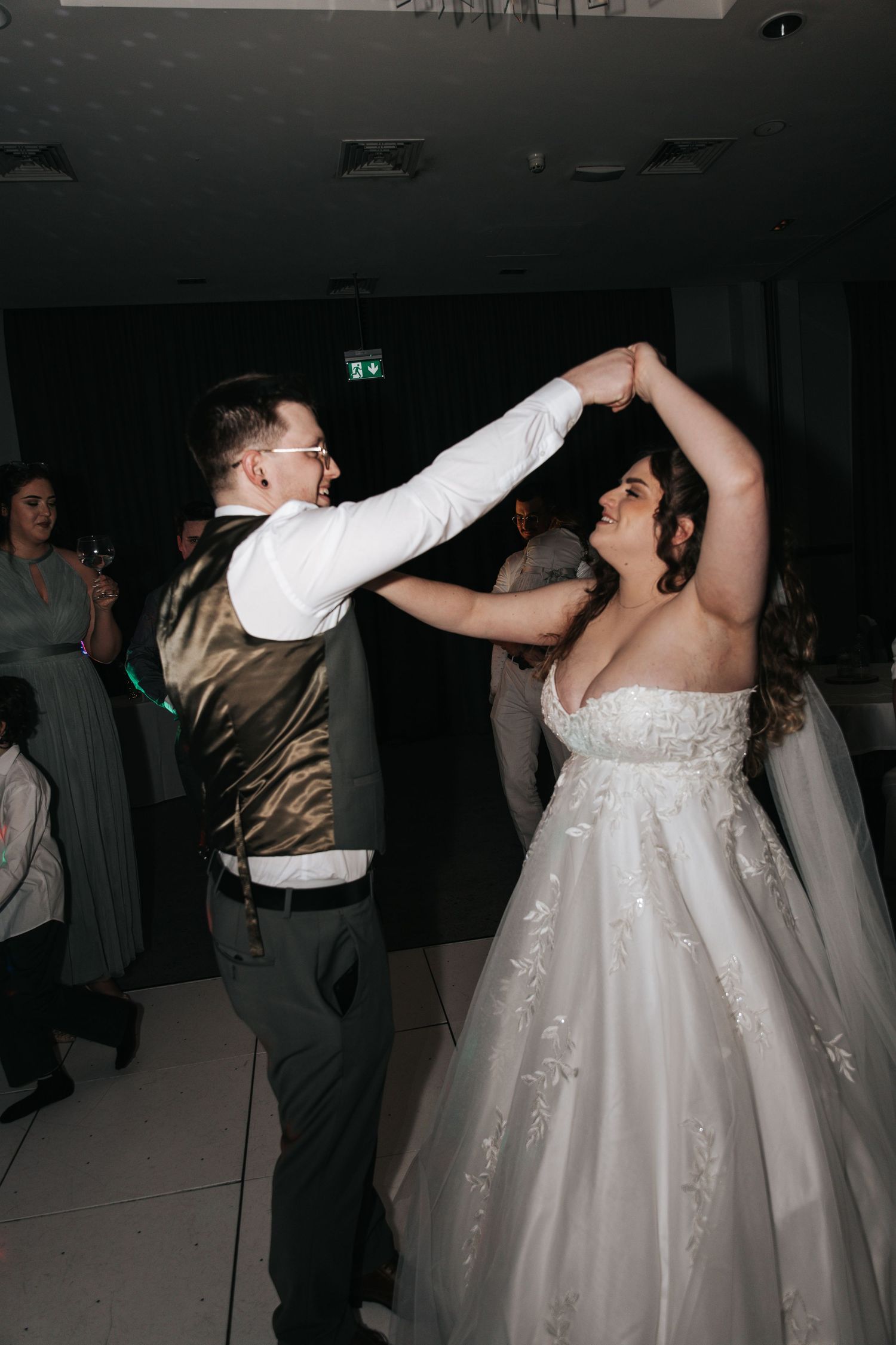 A couple dances together at their wedding reception, with the bride wearing a sparkly white gown and groom in vest and tie.