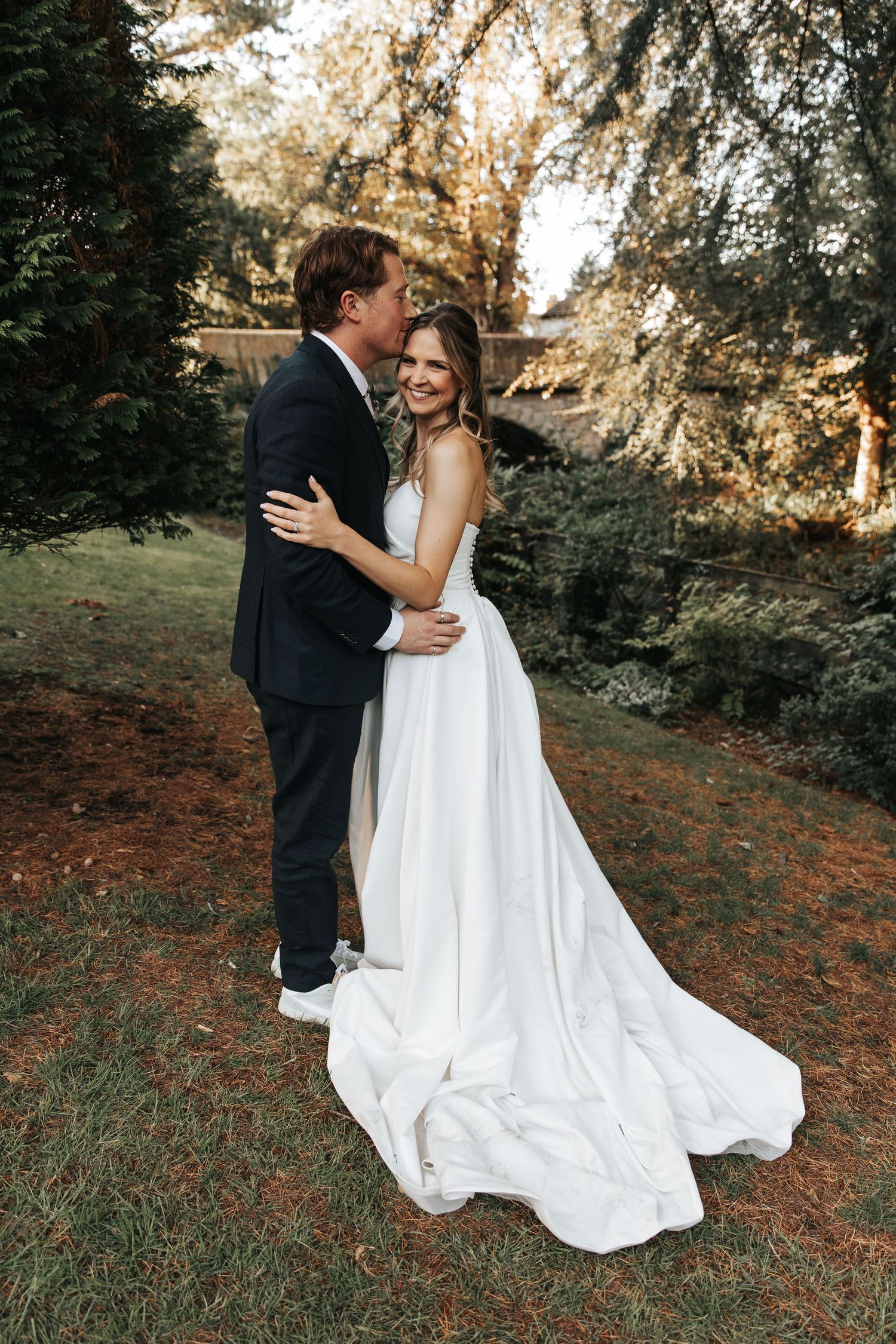 A bride in a white gown and groom in a dark suit embrace outdoors in a lush garden during their wedding day.