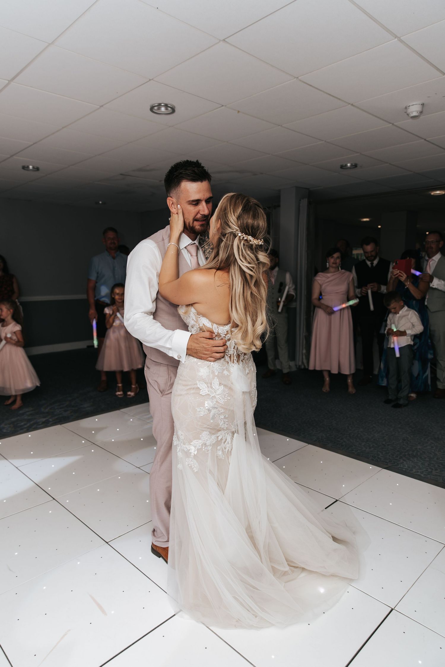 Bride and groom share their first dance on a white sparkle dance floor, guests watching in the background.