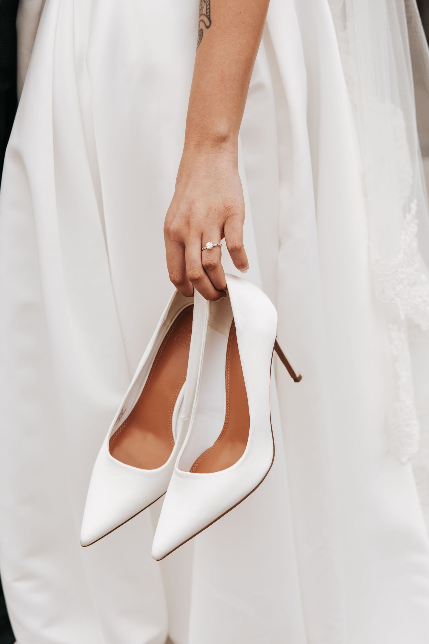 Close-up view of white satin bridal shoes with pointed toes and stiletto heels against flowing white wedding dress fabric.