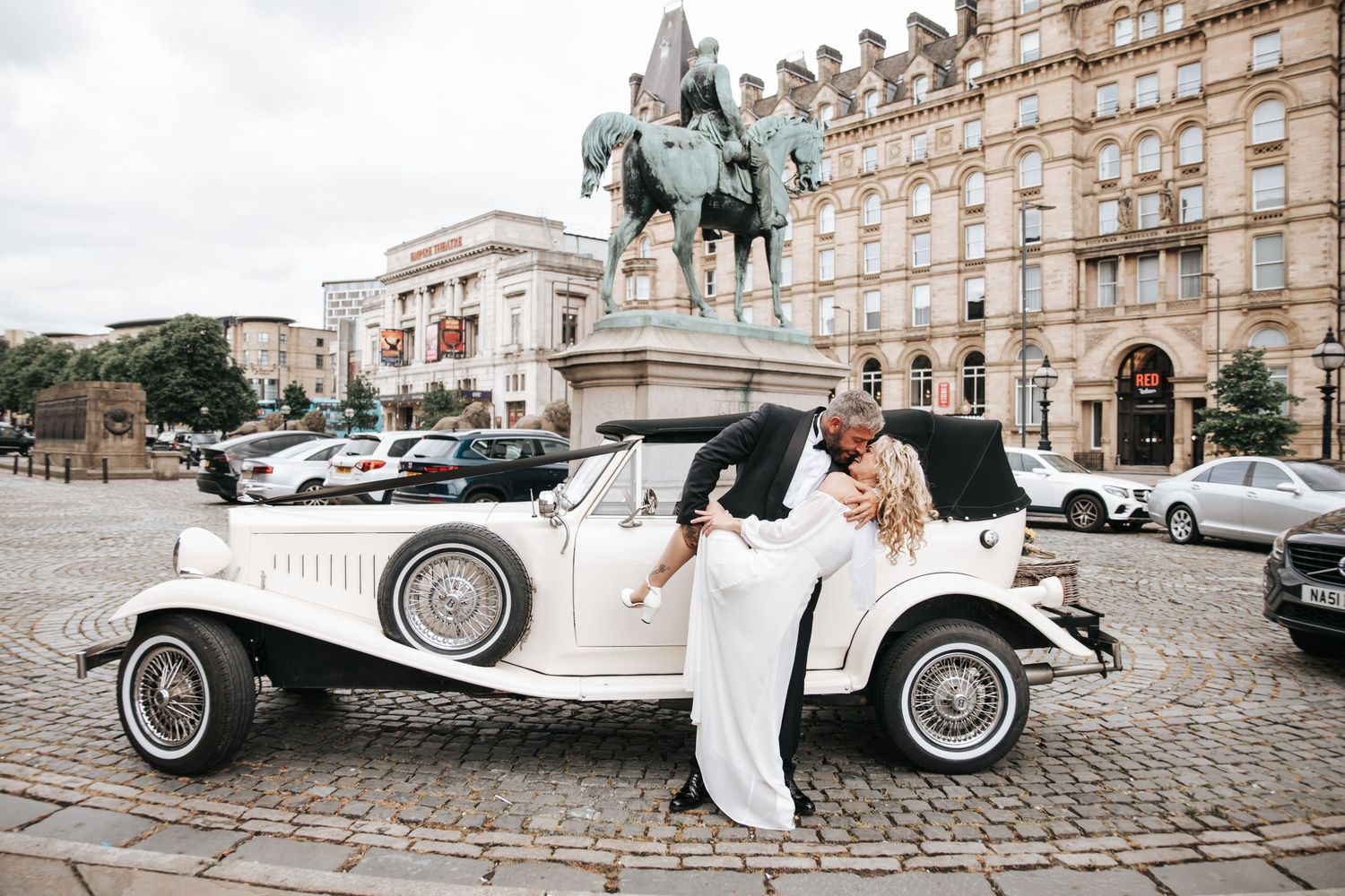 Bride and groom kissing beside a vintage white classic car on cobblestone square with equestrian statue backdrop.