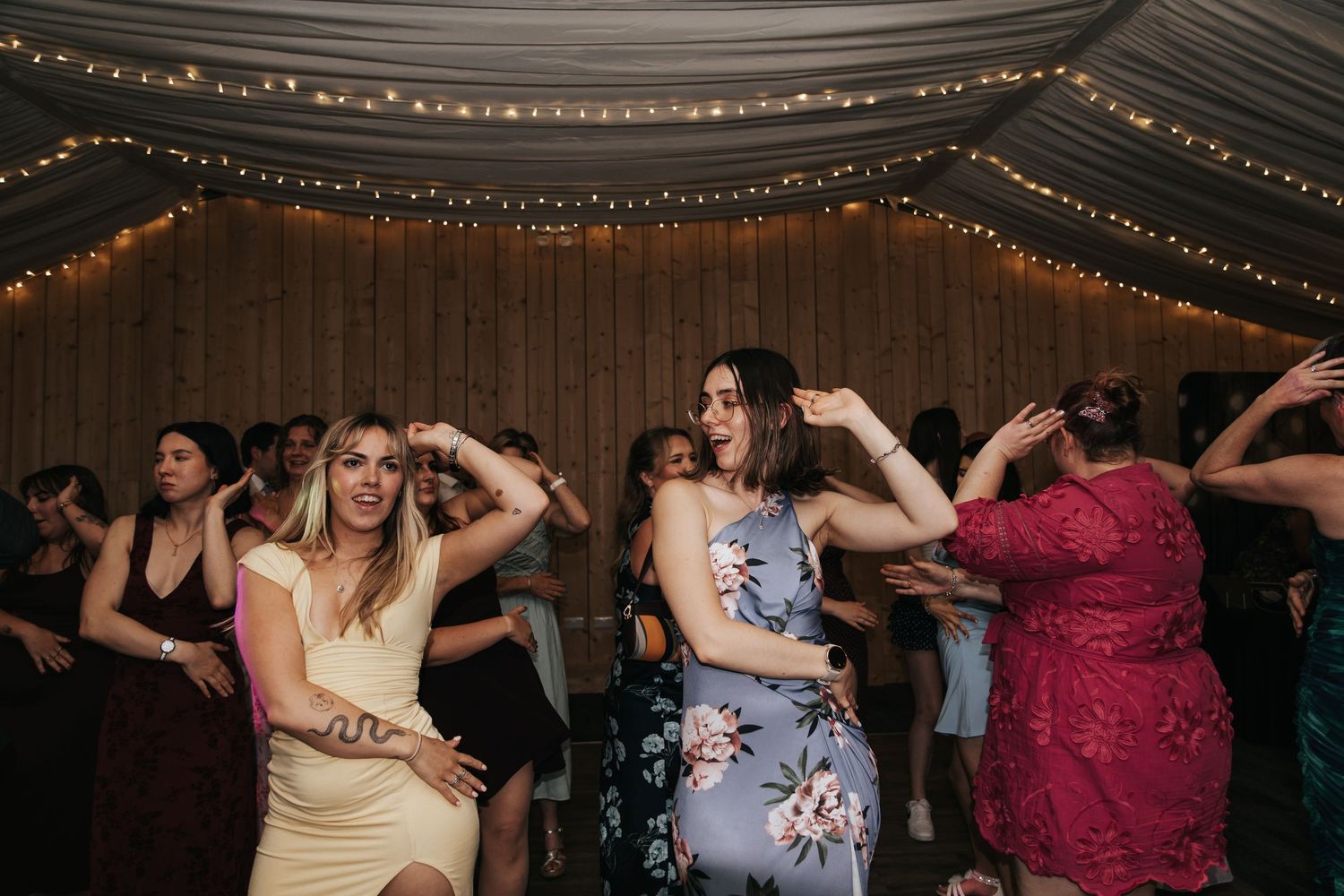 Guests dancing joyfully at a wedding reception with fairy lights and wooden backdrop.