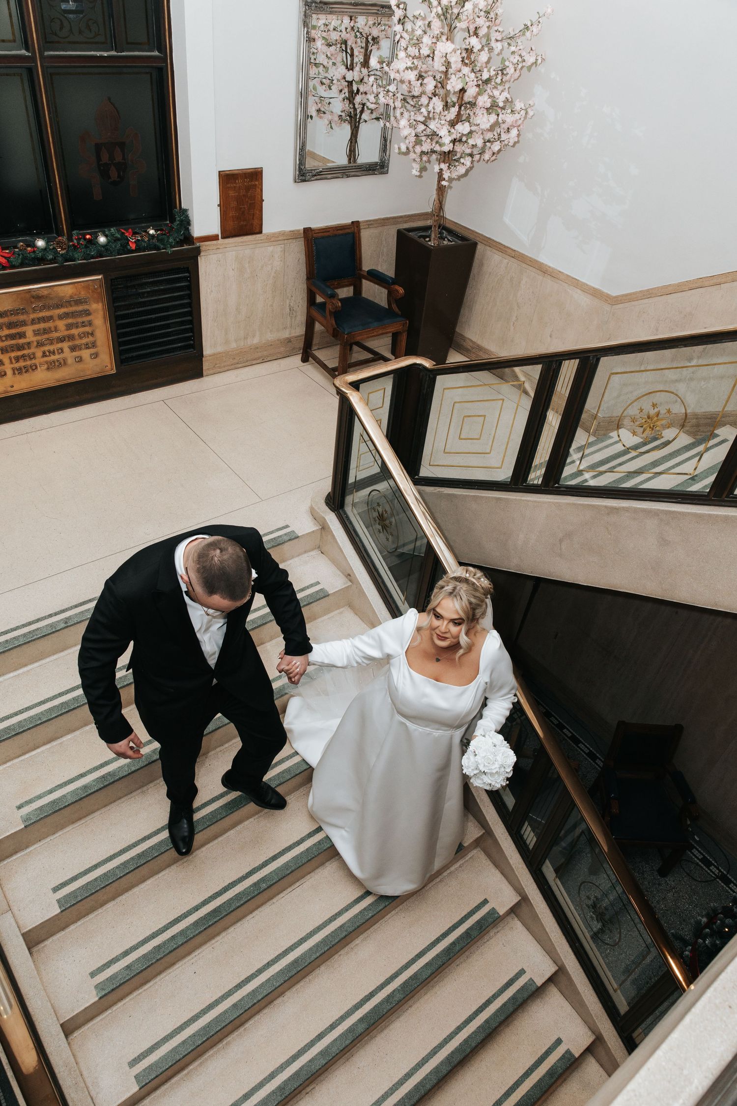 Bride and groom holding hands walking down elegant staircase in wedding attire at a formal venue.