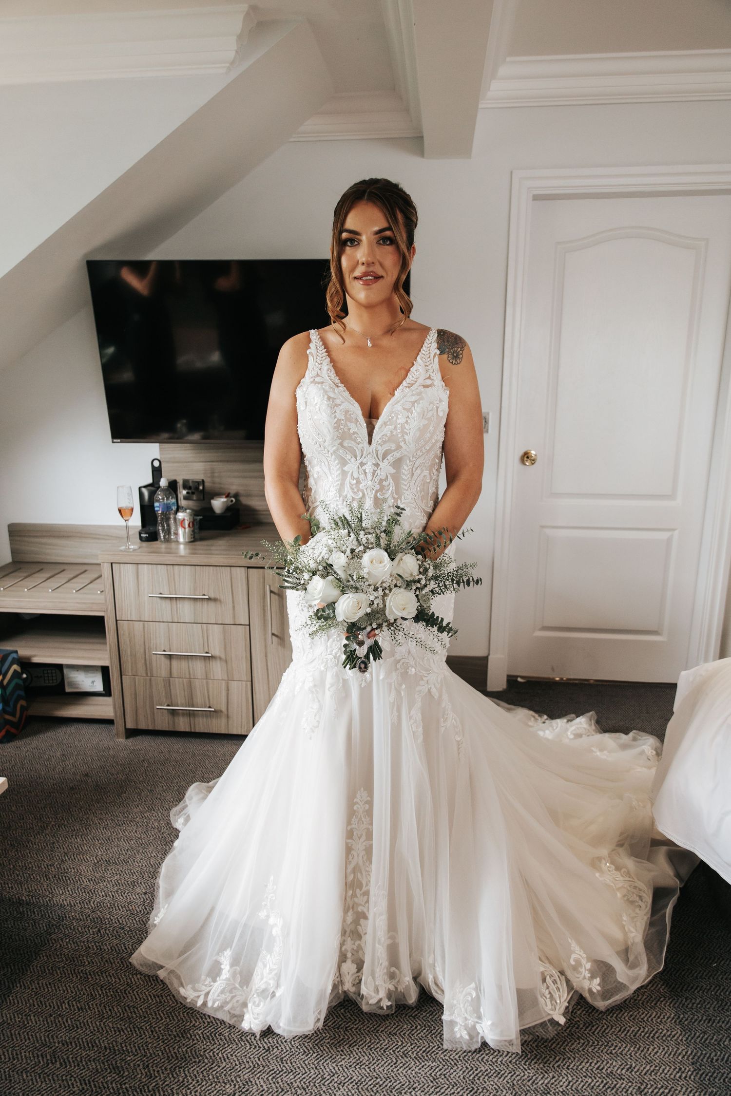 Bride poses in elegant lace mermaid wedding dress holding white bouquet in a bright hotel room.