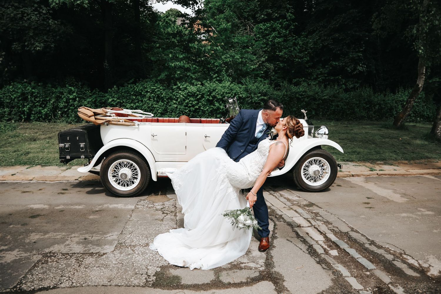 Bride and groom sharing a romantic dip kiss in front of a vintage white wedding car outdoors.