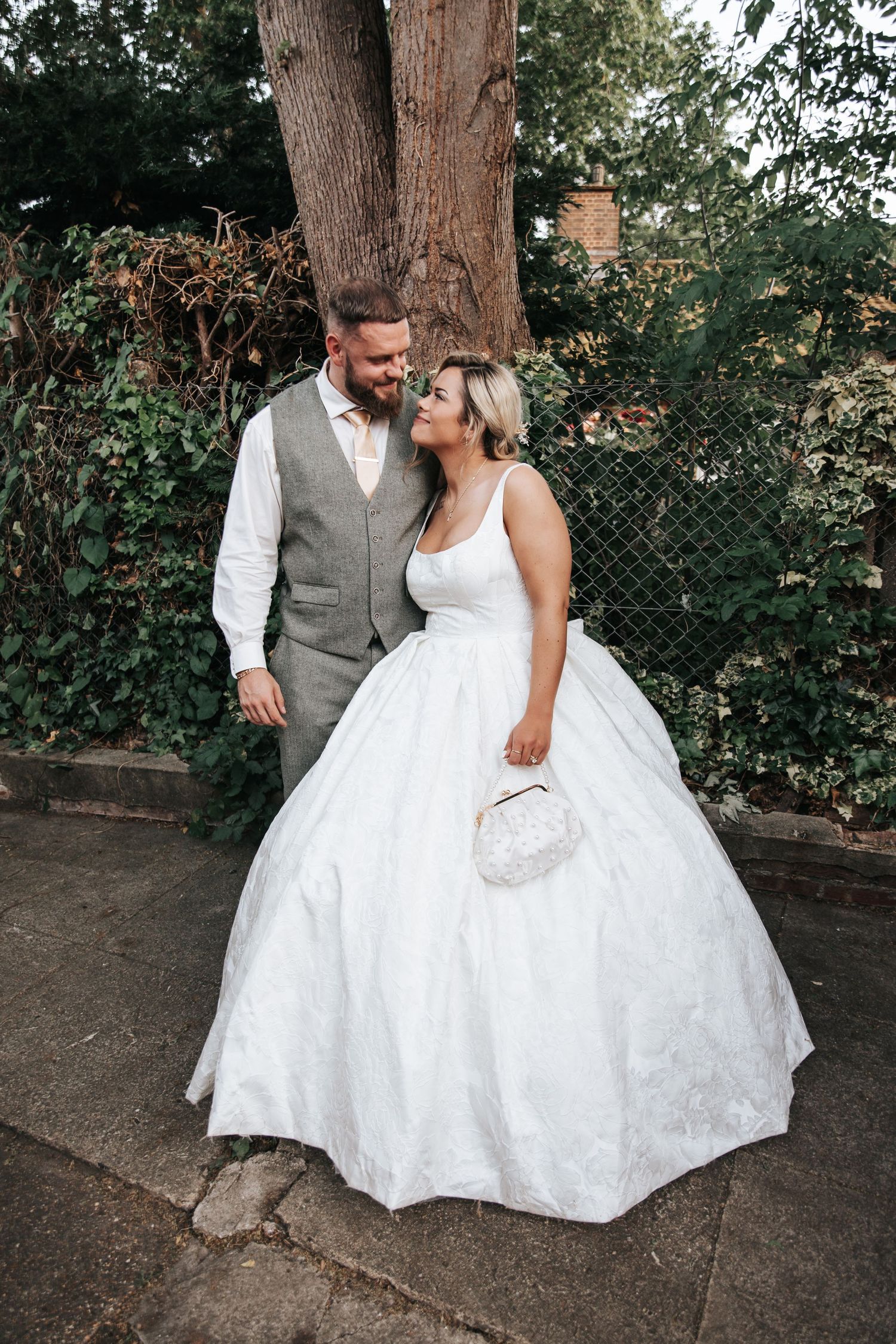 Bride in ball gown and groom in gray vest share a romantic moment by a tree outdoors.