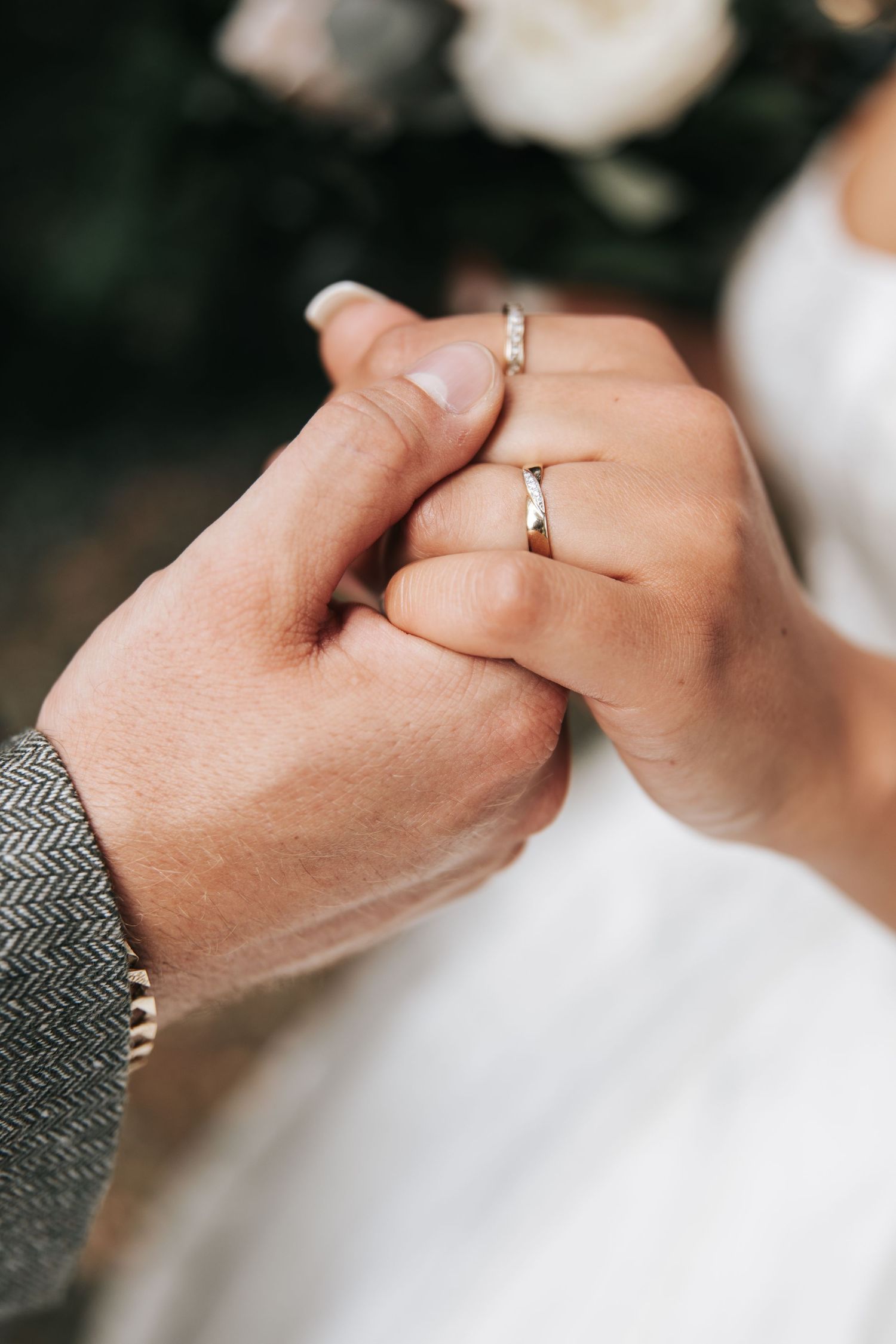 Newlywed couple holding hands showing matching silver wedding rings, bride in white dress and groom in gray suit.