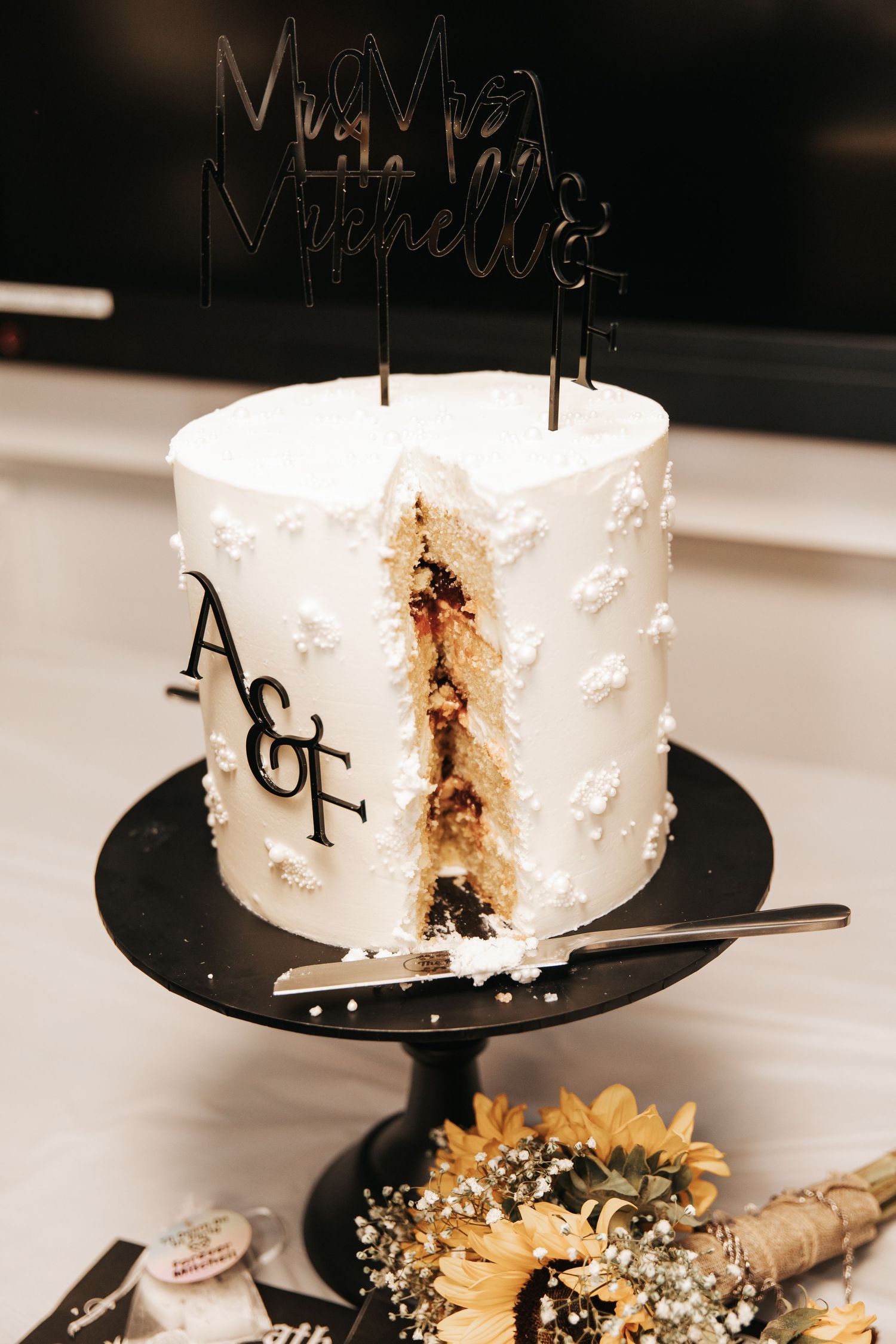 White wedding cake with A&F monogram, Mr & Mrs topper, floral details, sliced open on black stand with sunflowers nearby.
