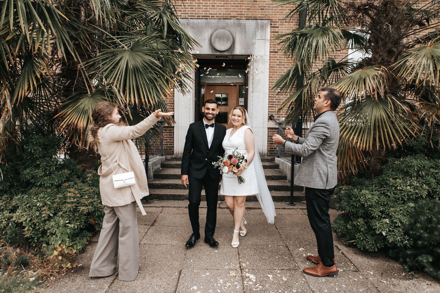 Wedding party gathering outside brick building with palm trees.