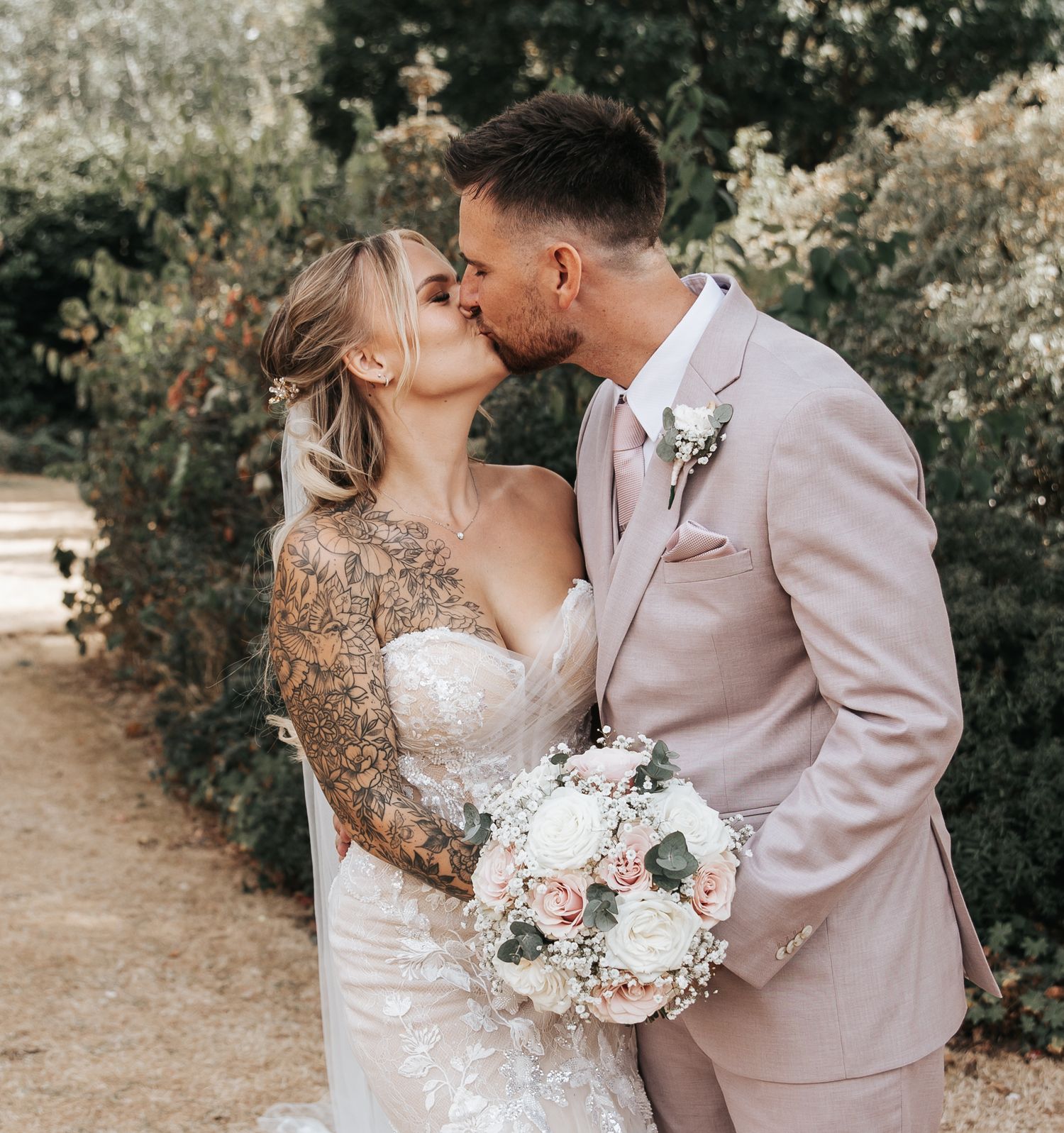 A bride and groom share a romantic kiss outdoors, she wears a lace gown with tattoos visible, holding a white floral bouquet.