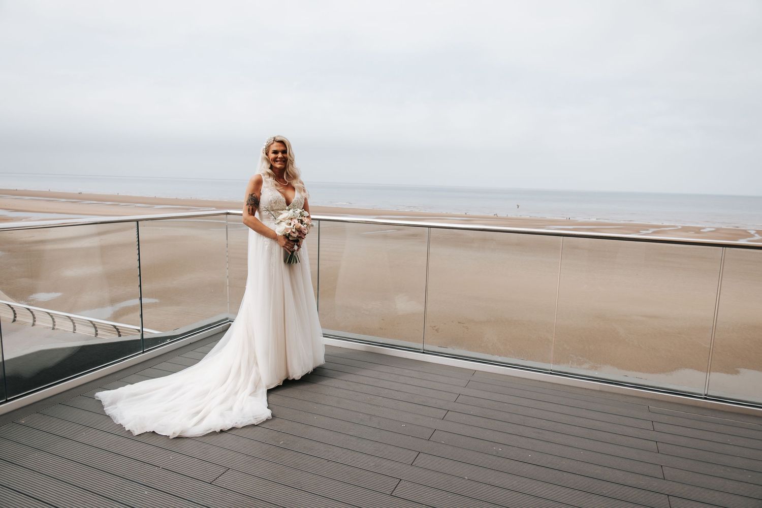 Bride in white lace gown stands on beachfront terrace holding bouquet with sandy shore and cloudy sky backdrop.