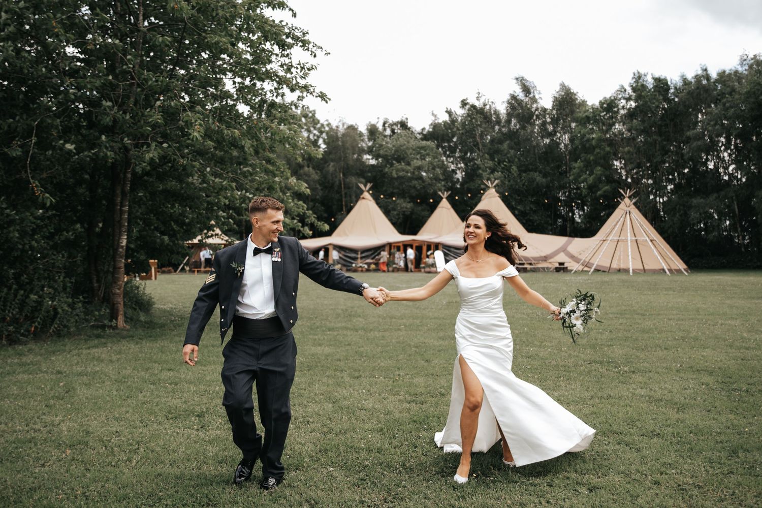 A joyful bride and groom hold hands running across green lawn with rustic tipi wedding venue in background.