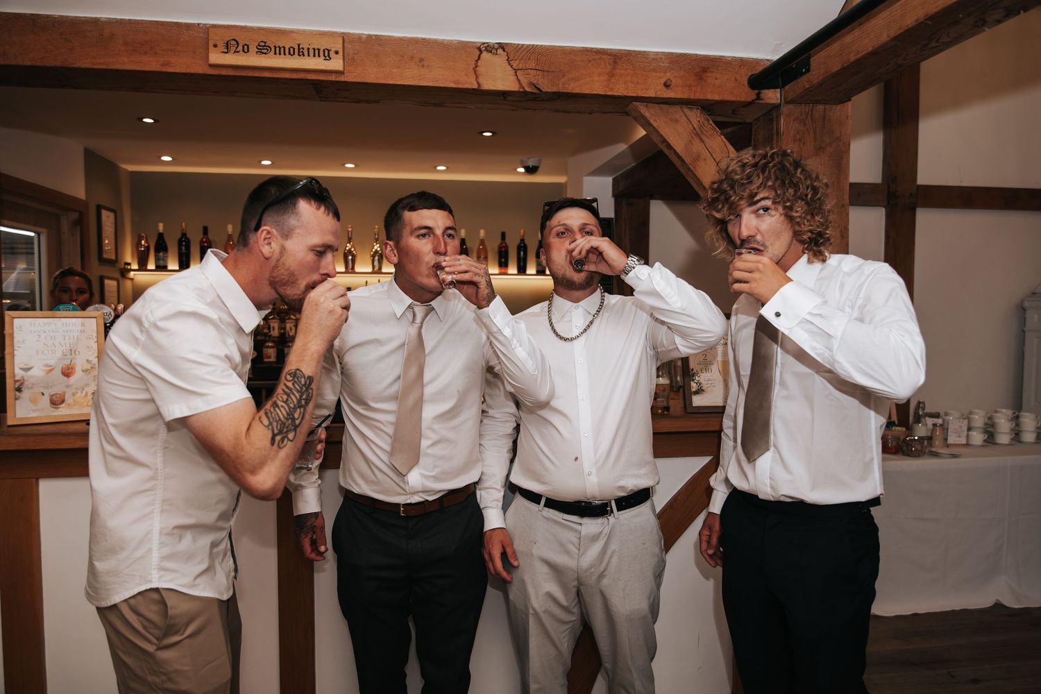 Four groomsmen in white shirts taking shots together at a wedding reception bar.