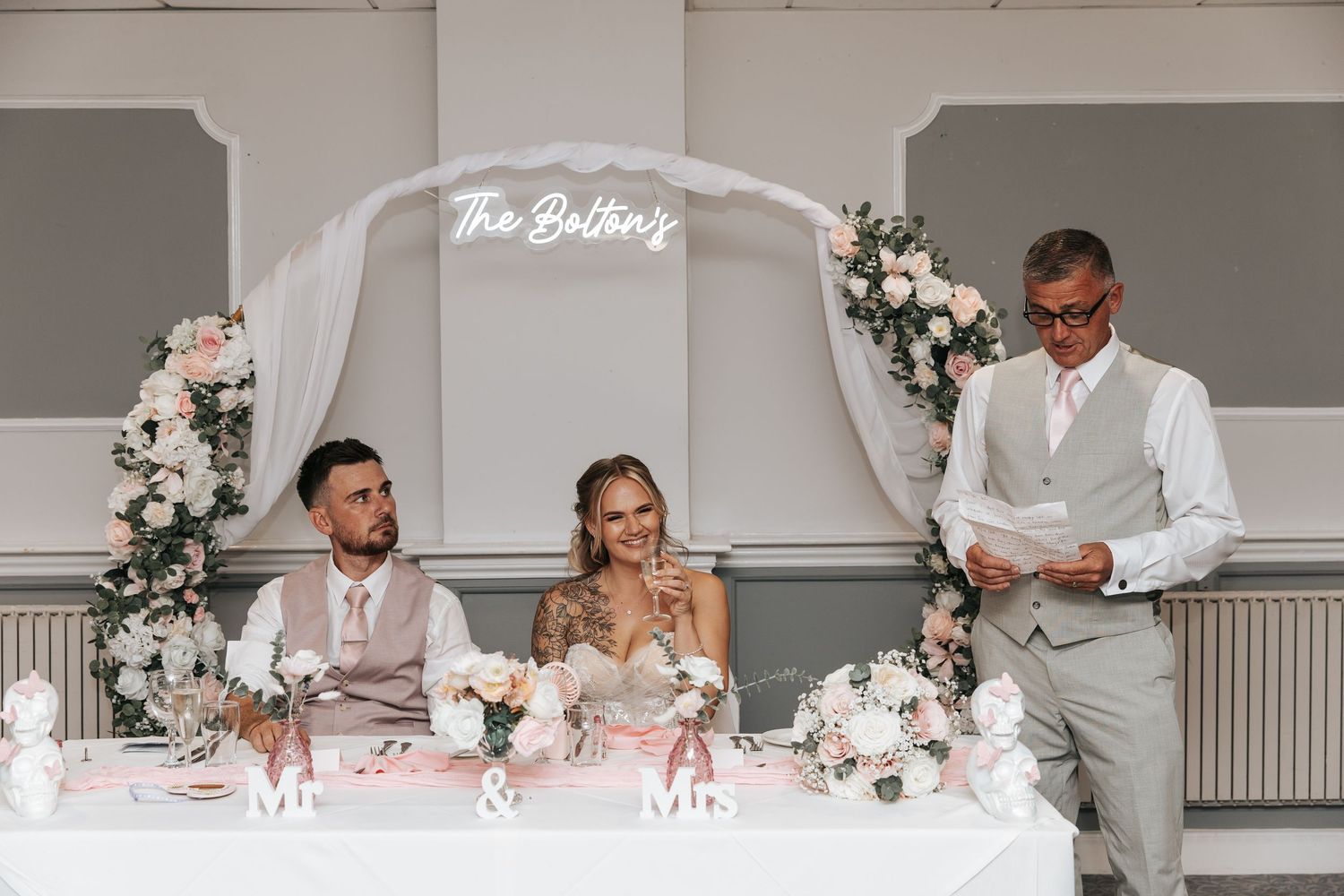 Bride and groom seated at wedding reception sweetheart table with floral arch and The Boltons neon sign.