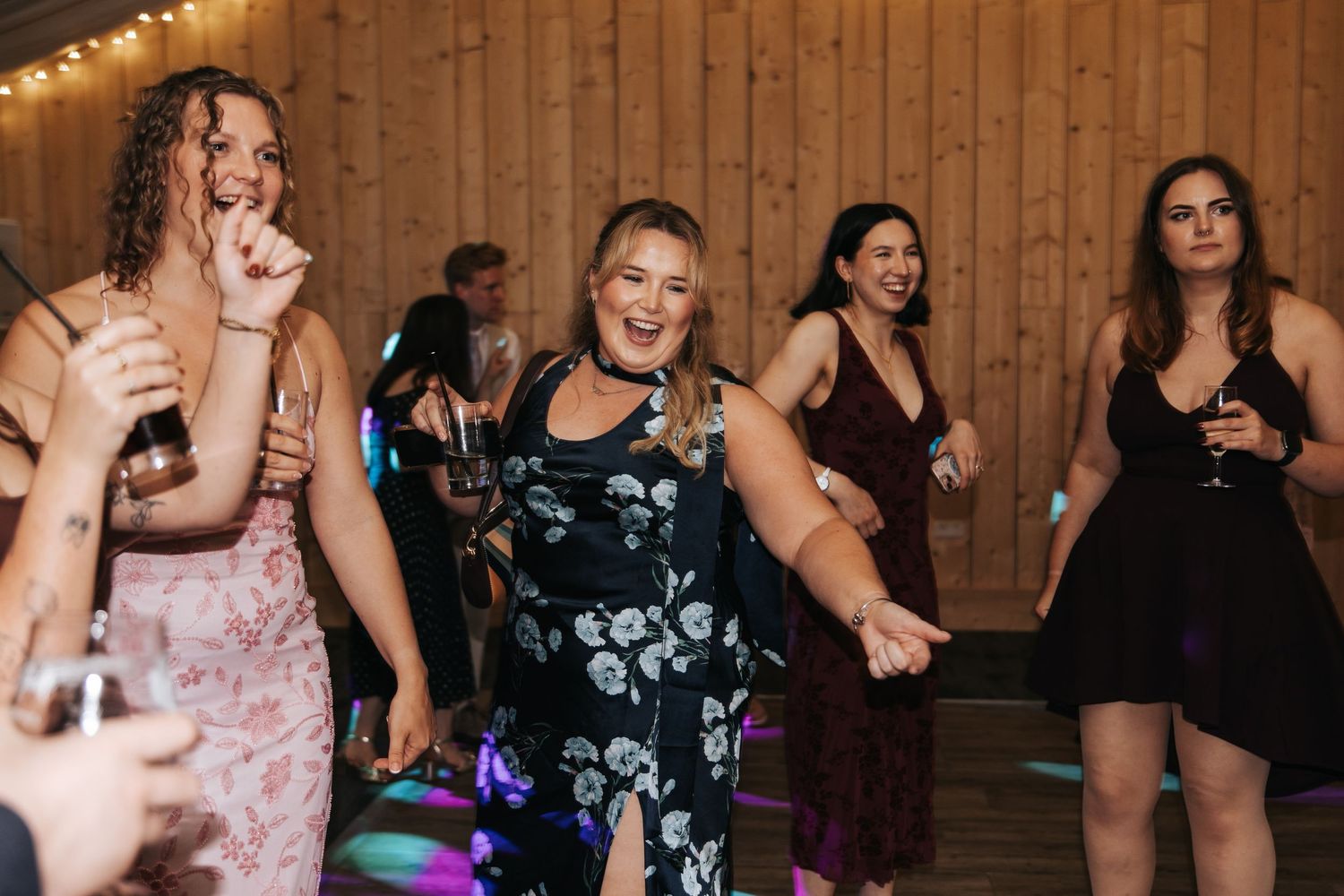 Guests in elegant dresses laughing and dancing at a wedding reception with colorful dance floor lights.