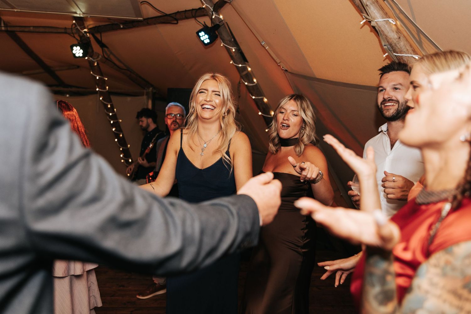 Guests dancing and laughing joyfully at an elegant wedding reception under a tent with string lights.