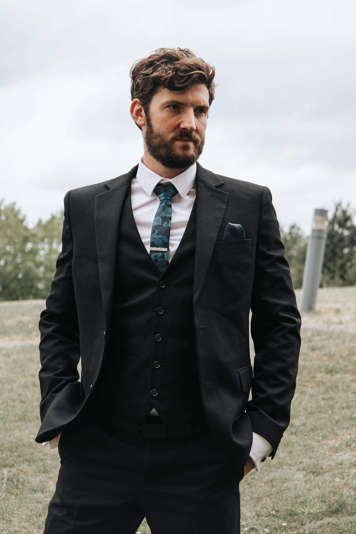 A person in a three-piece suit stands outdoors on a grassy hillside.