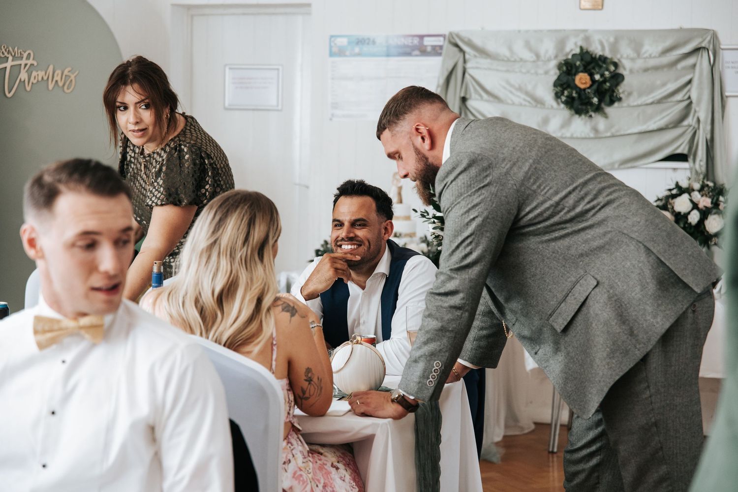 Guests socializing at a wedding reception, with a smiling groom in a gray suit greeting seated attendees.