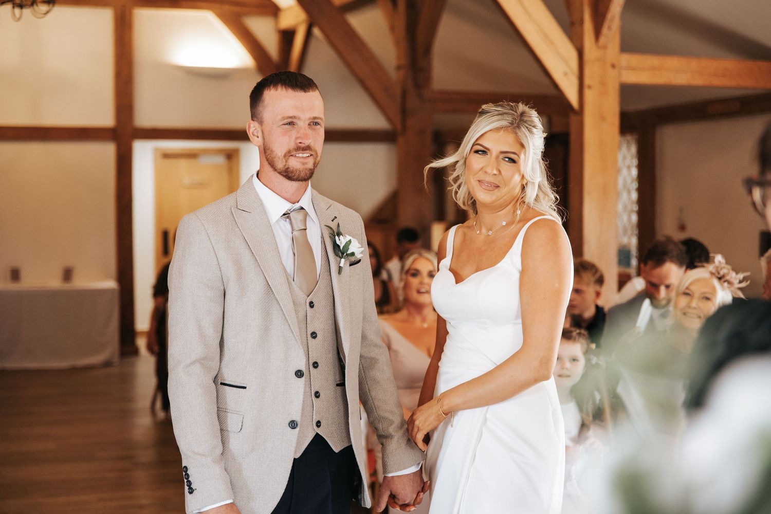A smiling bride and groom hold hands at their rustic barn wedding reception, she in white gown, he in beige suit.
