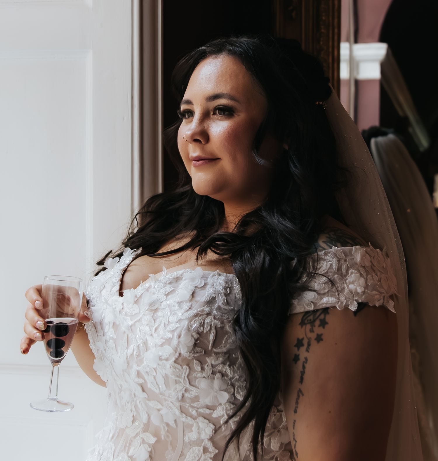 Elegant bride in lace wedding dress holding champagne glass by window.