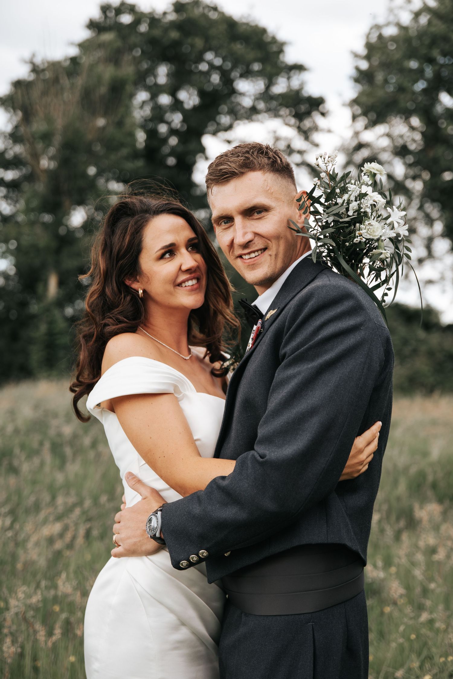 Smiling bride and groom embrace in a field, she in a white off-shoulder gown, he in a dark suit with floral boutonniere.