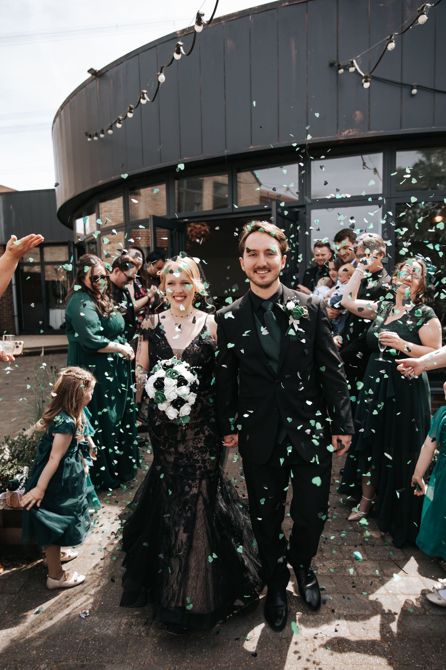 Bride in black lace gown and groom exit wedding ceremony through green confetti shower with guests cheering.