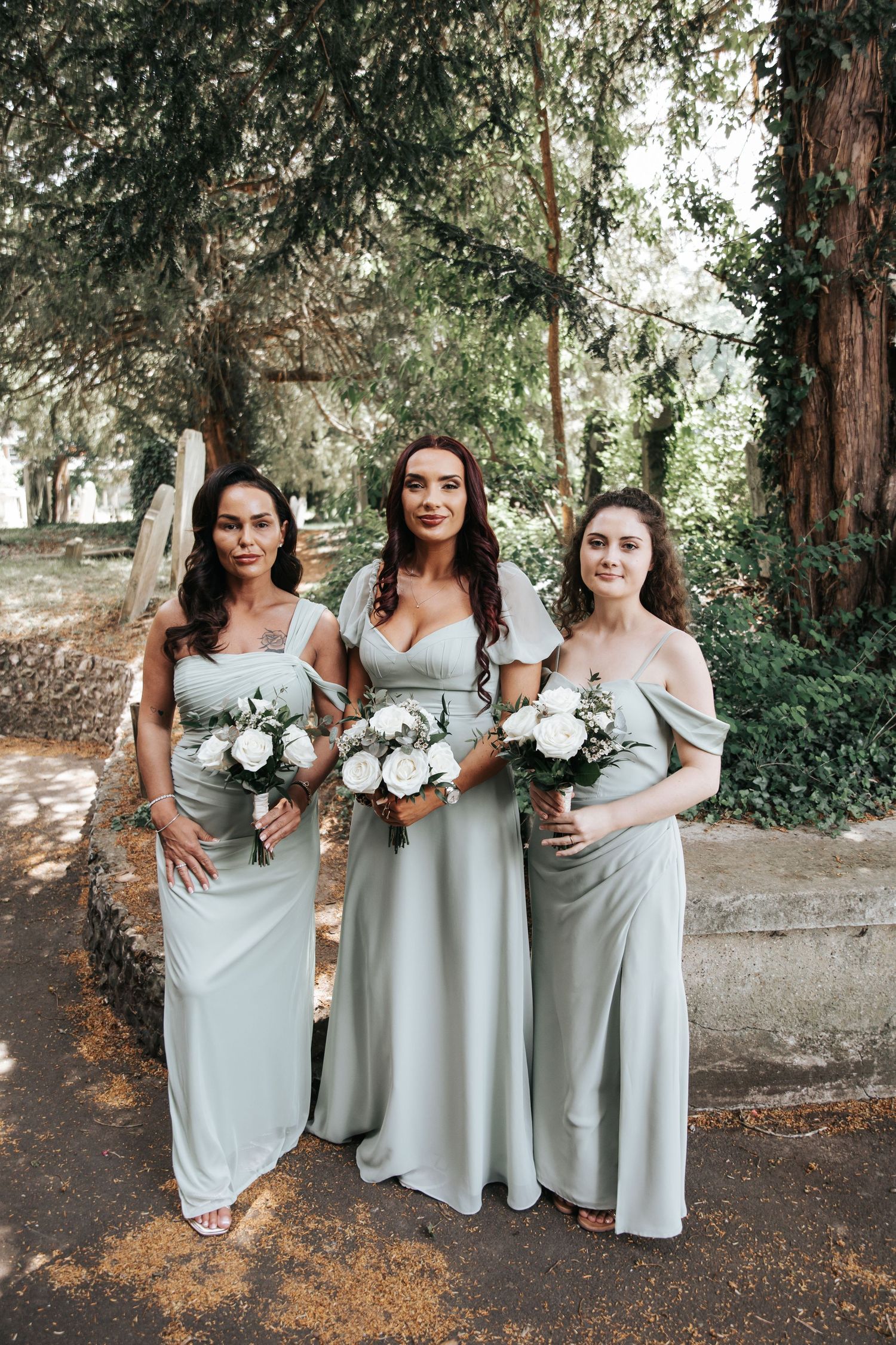 Three bridesmaids in sage green gowns holding white bouquets pose outdoors under lush trees at a wedding.