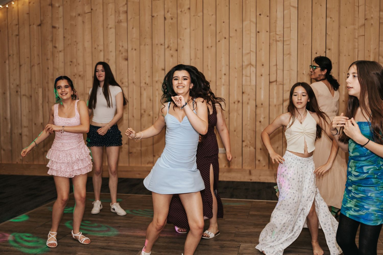 Group of young teenage girls dancing energetically at a party in a wood-paneled venue.