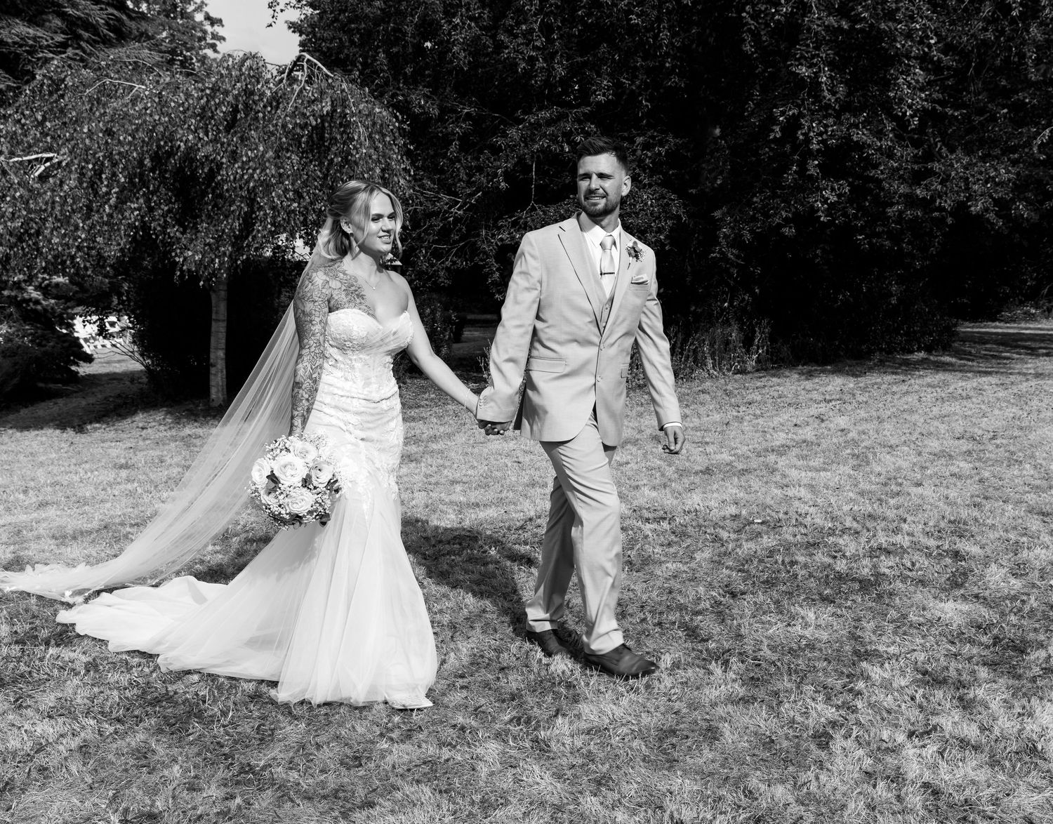 Black and white photo of a bride in a white gown and groom in a tan suit holding hands on a grassy lawn.