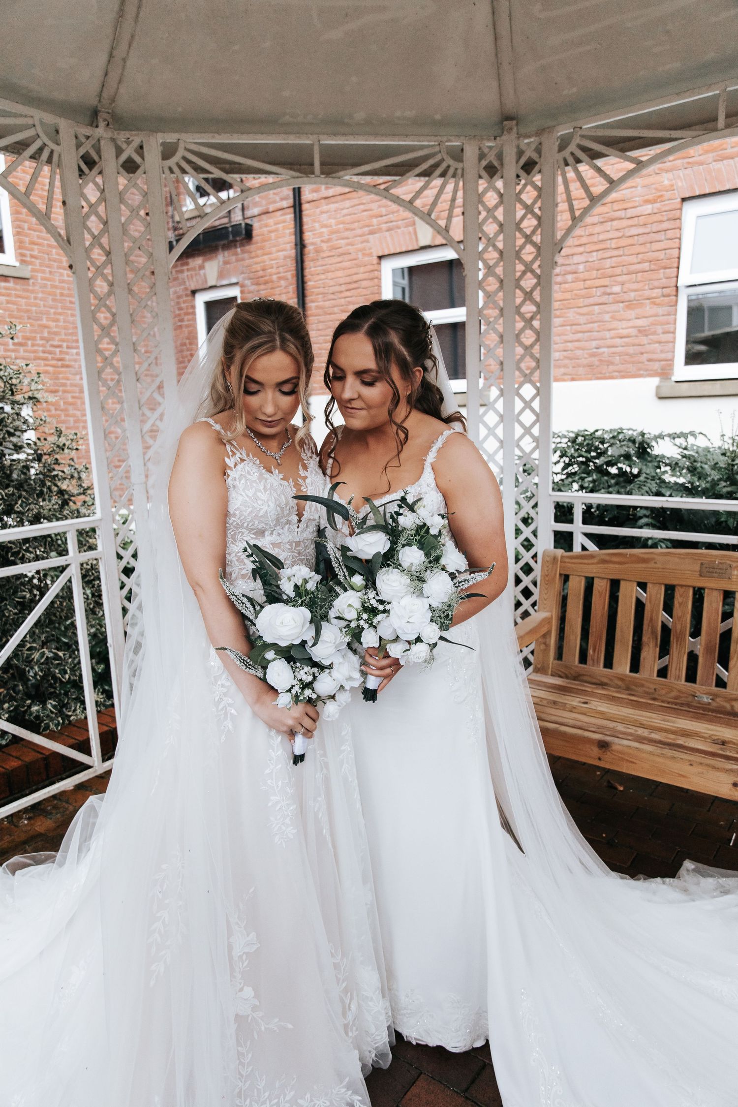 Brides share intimate moment under gazebo with brick building backdrop.
