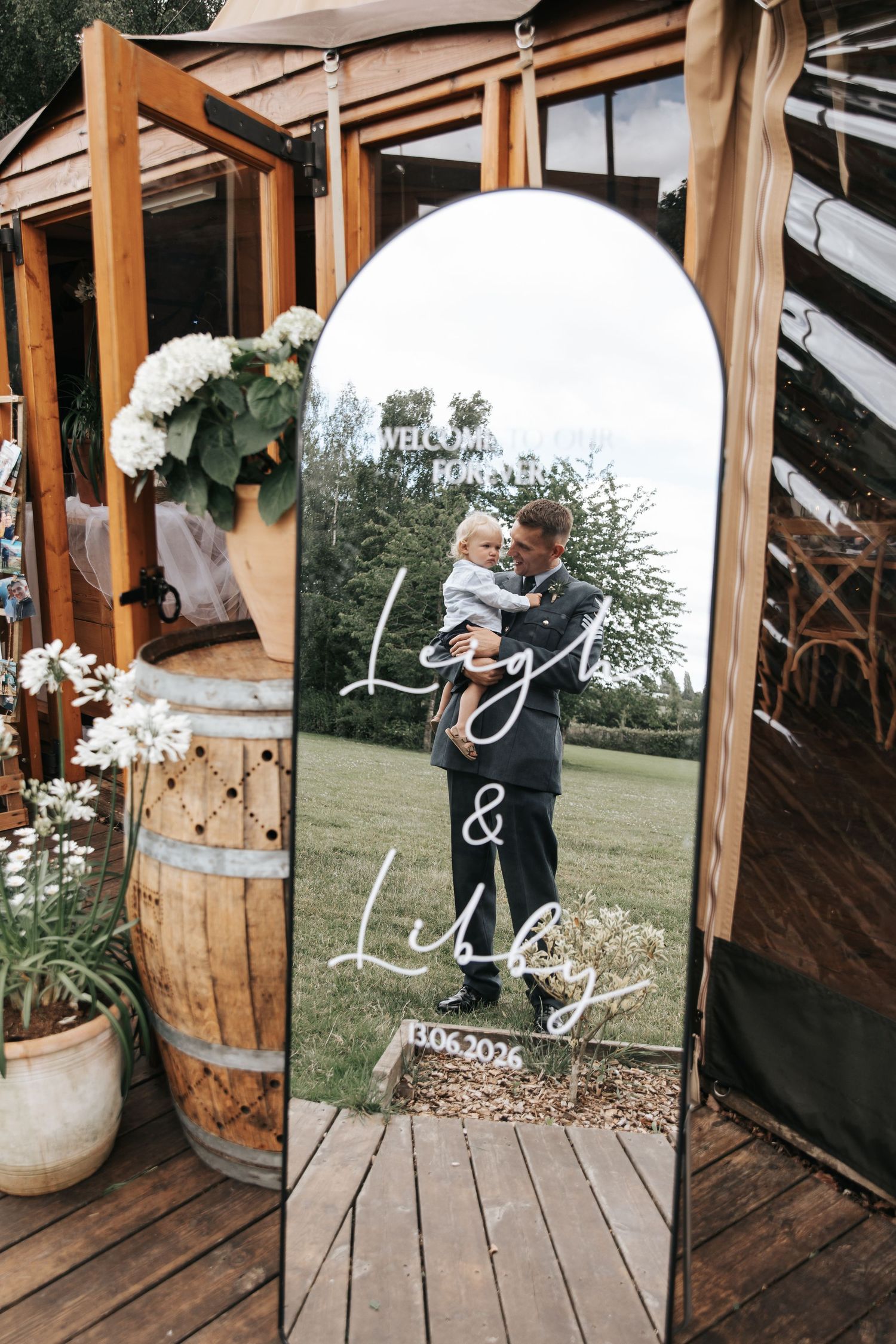 Wedding welcome mirror sign for Leigh and Libby with elegant script lettering, displayed outdoors beside a rustic wooden barrel.