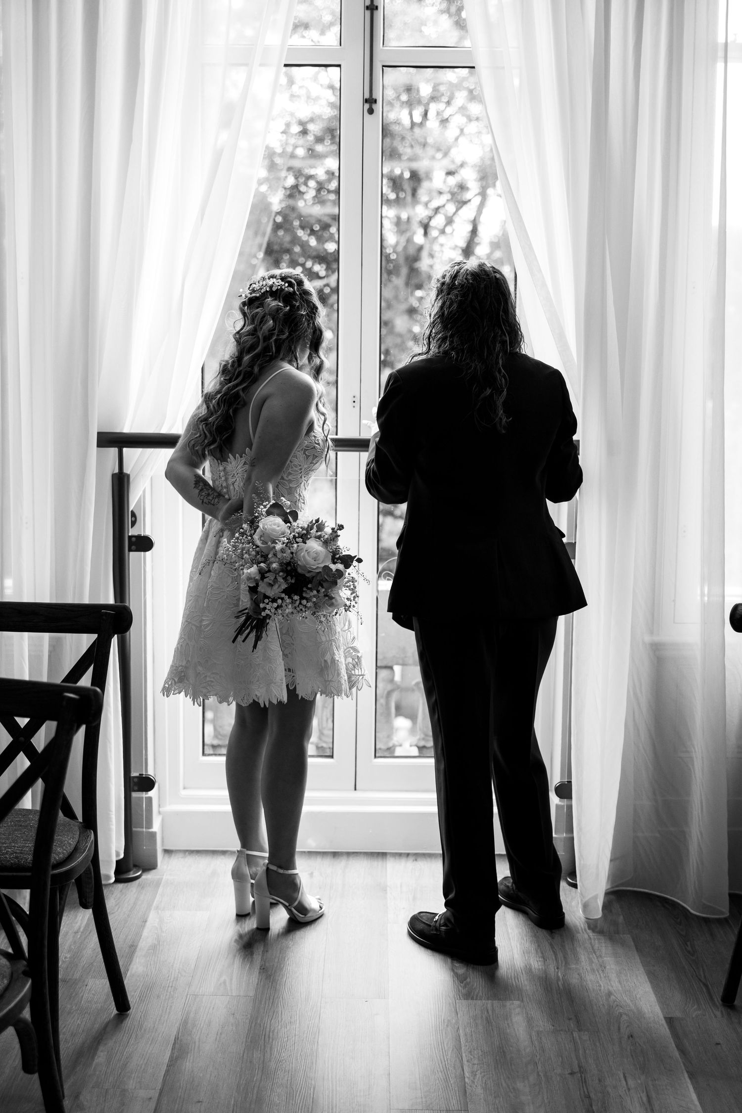 Black and white photo of a bride in short lace dress holding bouquet standing with guest by curtained window.