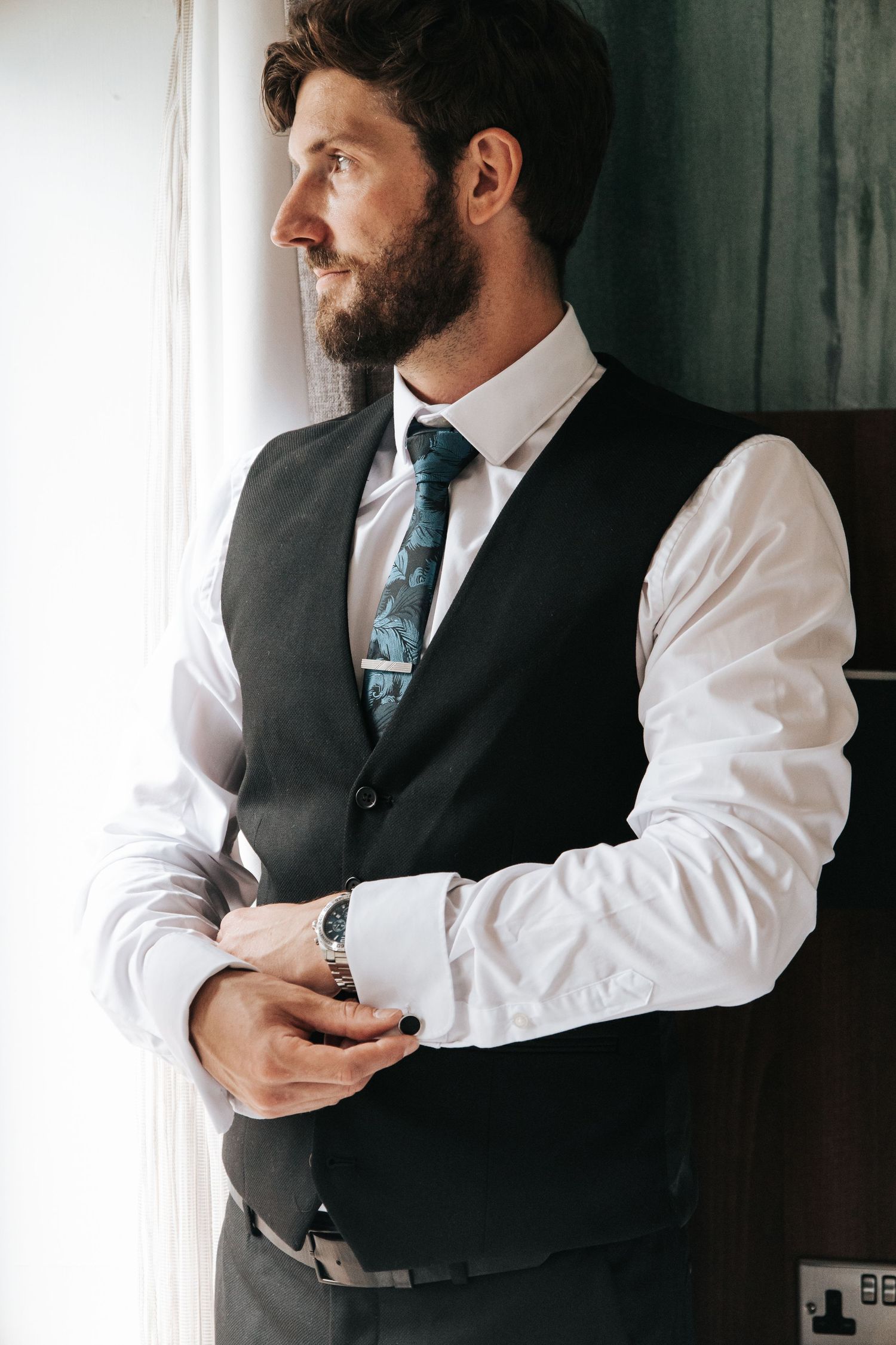 Man in formal vest and white shirt adjusting cuffs by window.
