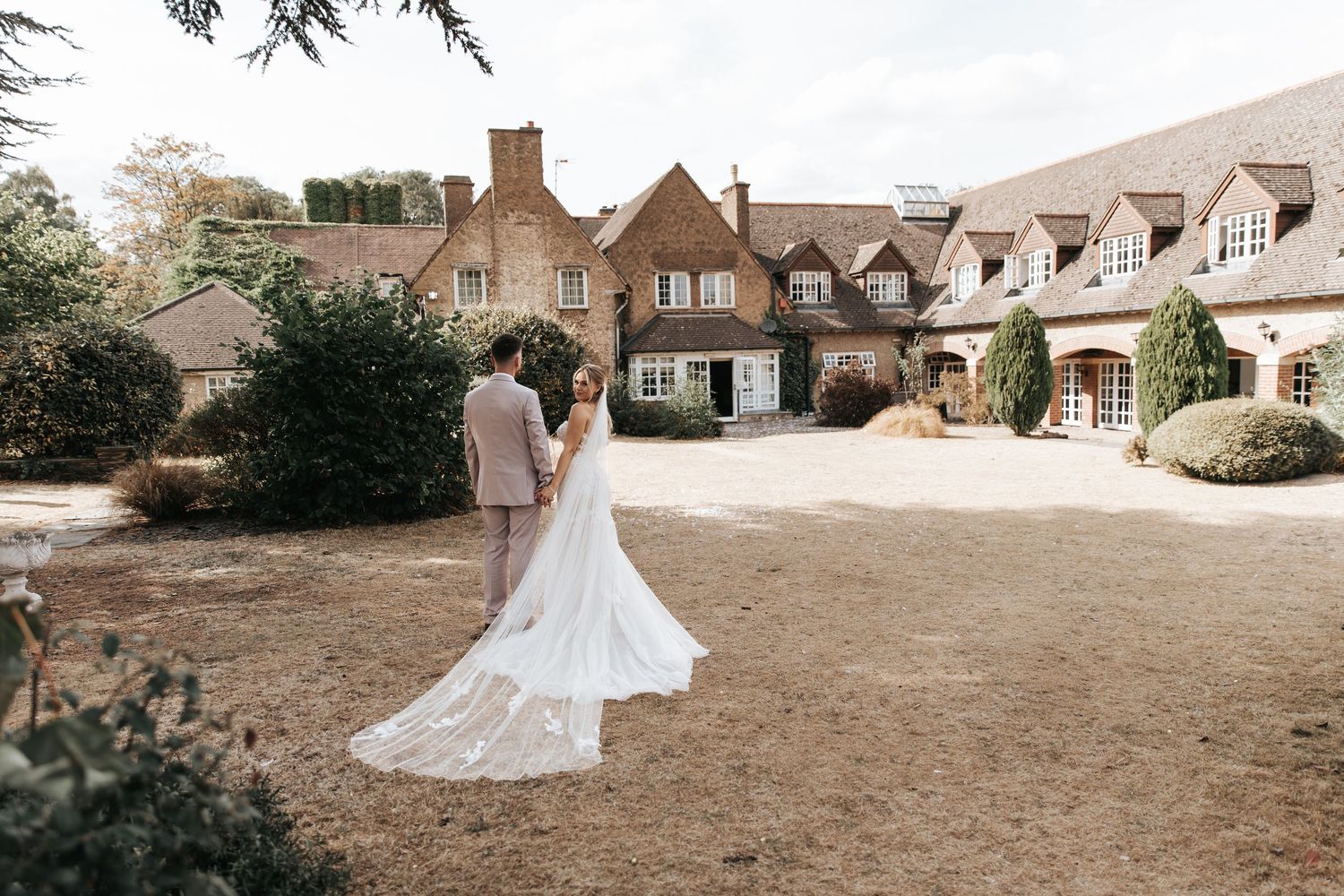 Bride and groom walk hand in hand toward a charming English manor house on their wedding day.