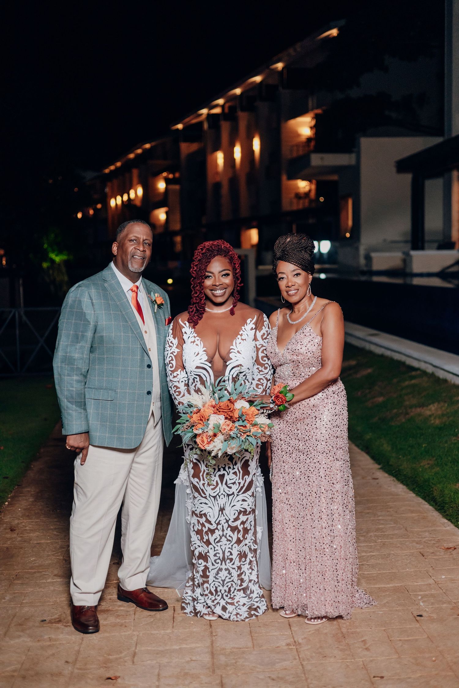Four festive guests in elegant attire pose at a formal evening event on a garden path lit by decorative string lights.