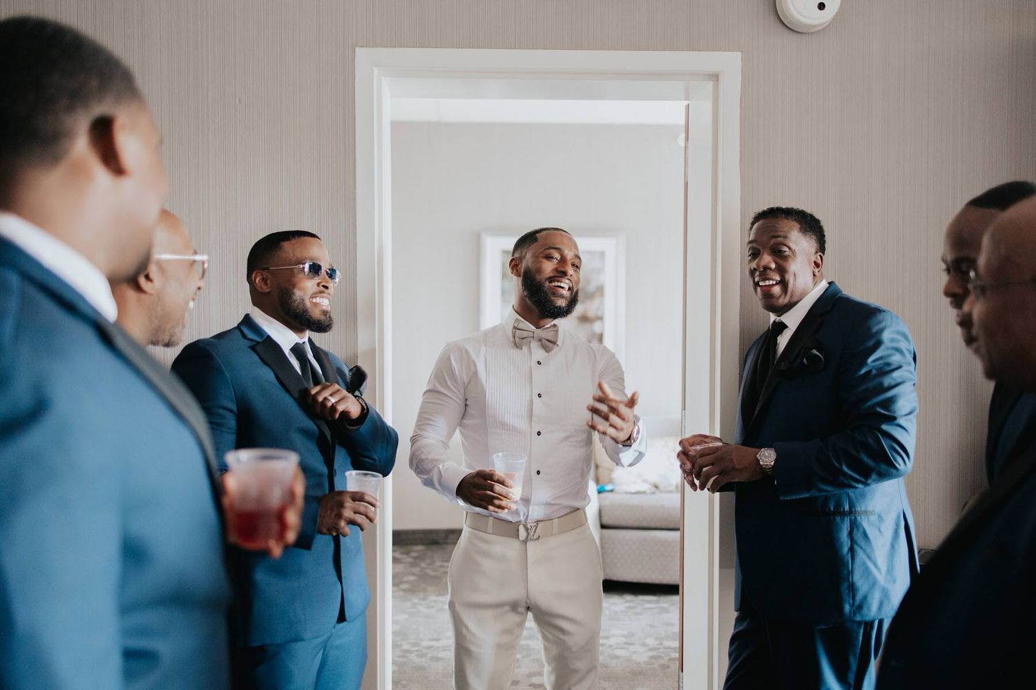 A group of men in suits and formal attire share a lighthearted moment while getting ready in a room.