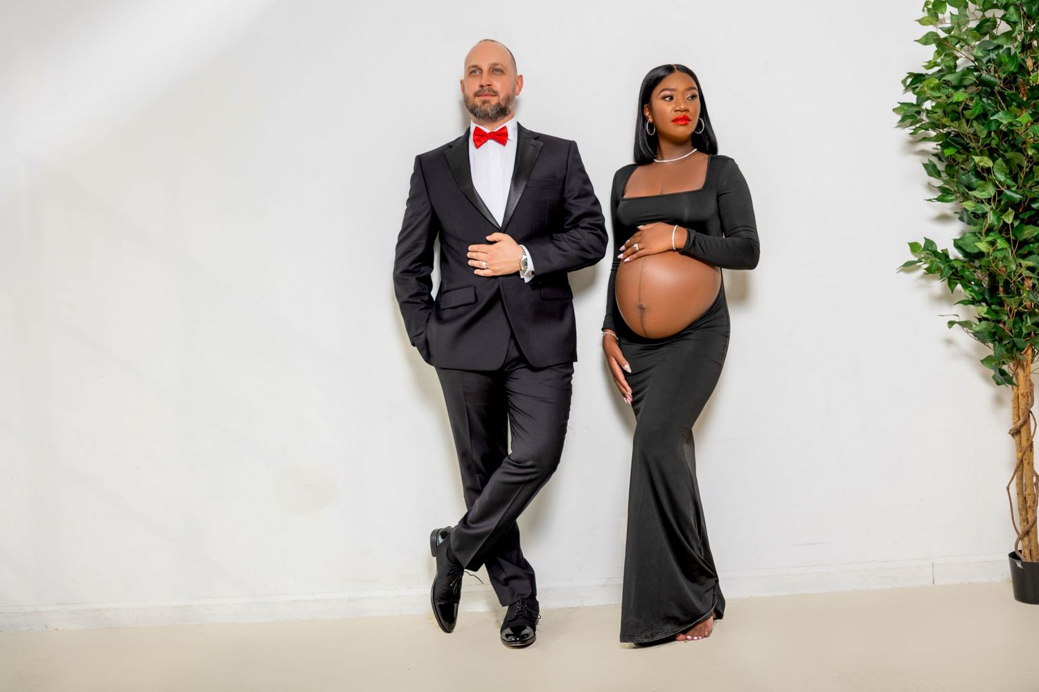 Maternity photo shoot featuring an elegant couple in black formal attire posing against a white backdrop with greenery.