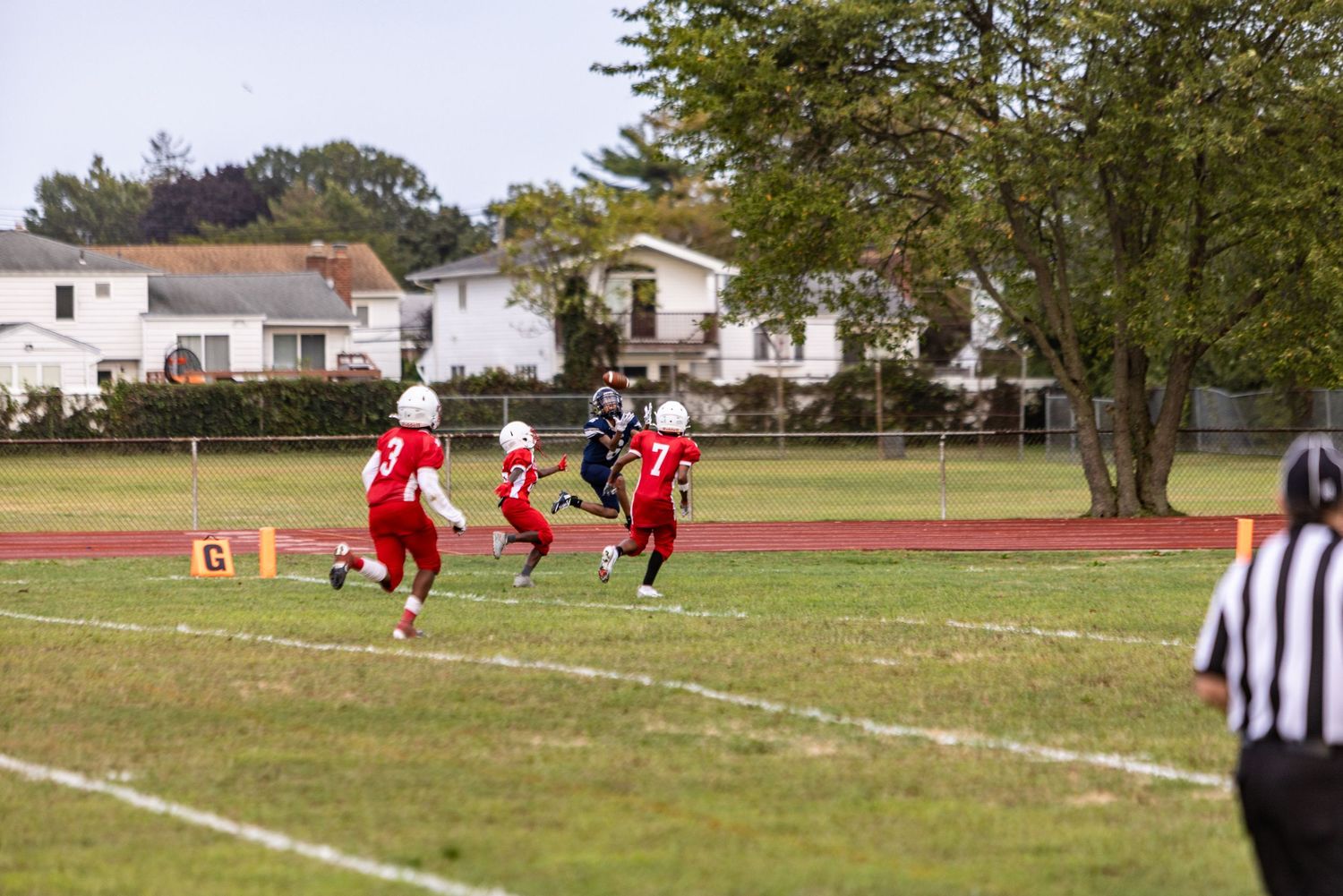High school football players run drills on a practice field while coaches and officials observe the training session.