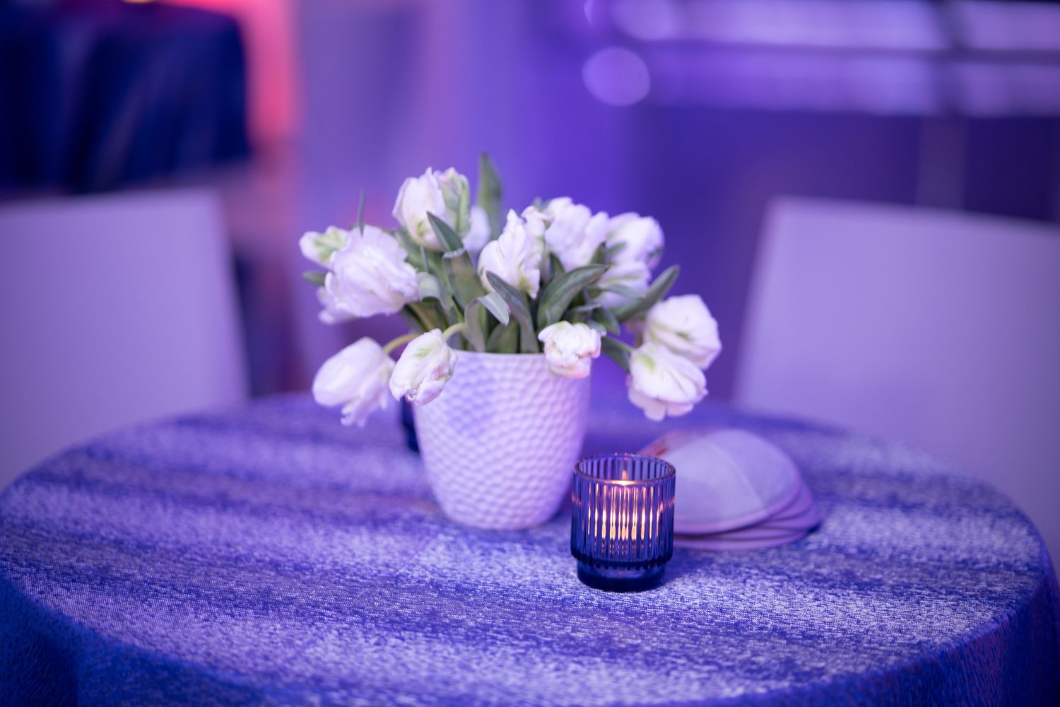 White tulips in a textured vase with candle on purple tablecloth under ambient lighting.