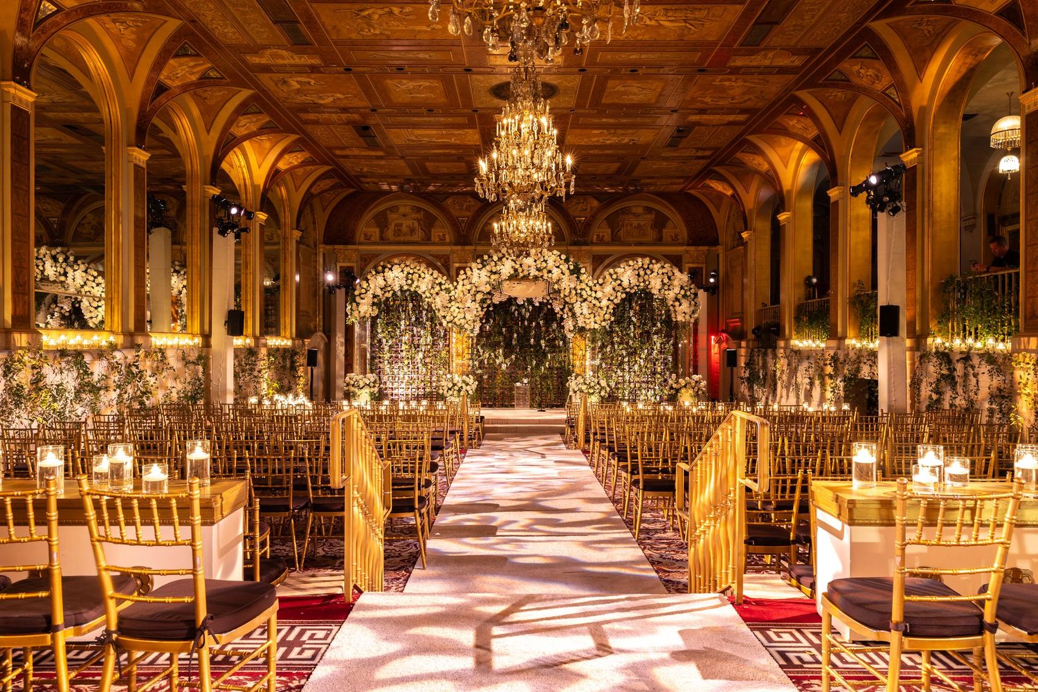 Elegant ballroom with crystal chandeliers and golden decor features rows of tables set for a luxurious wedding reception.