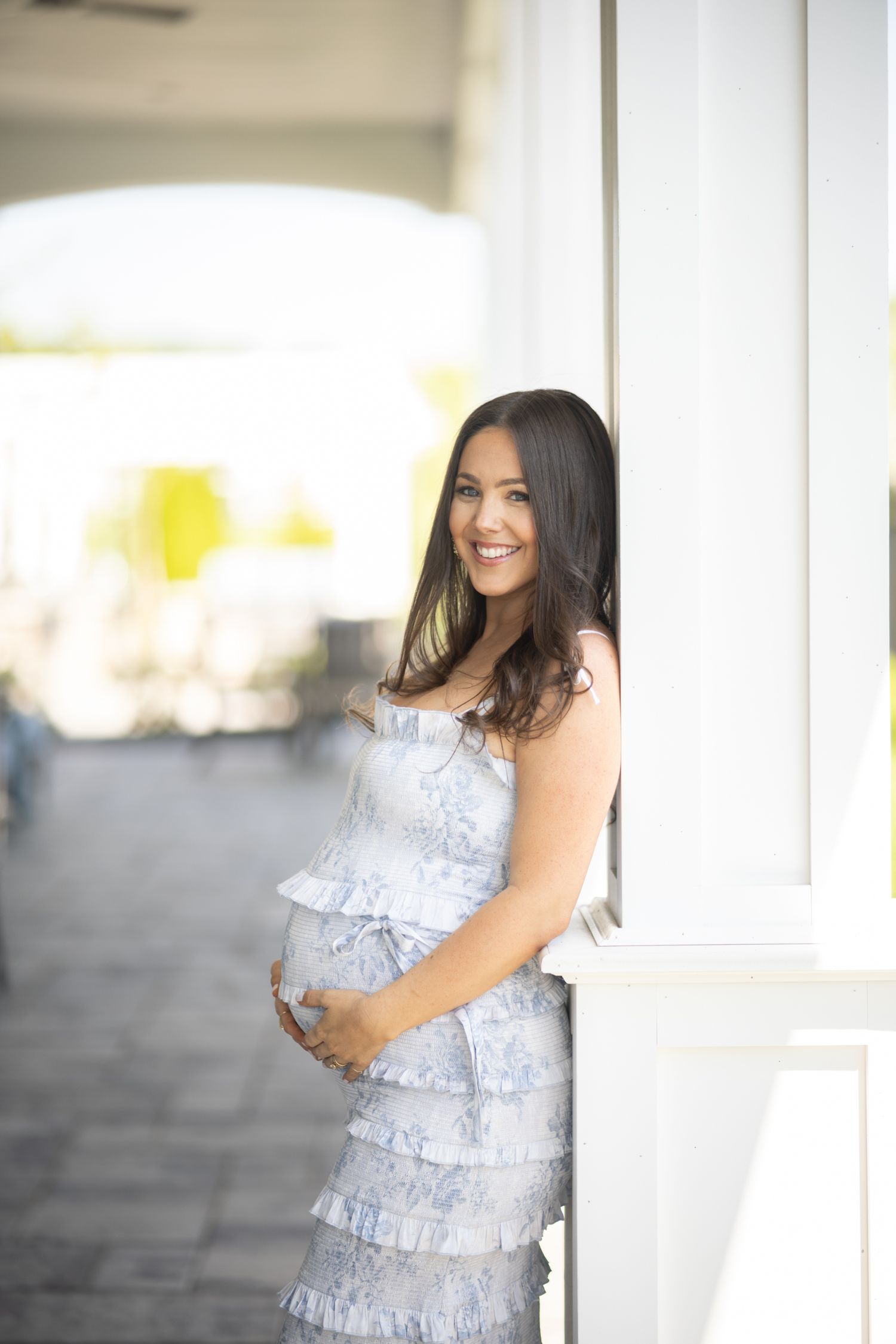 Expectant woman in a light blue ruffled dress poses against a white column outdoors.
