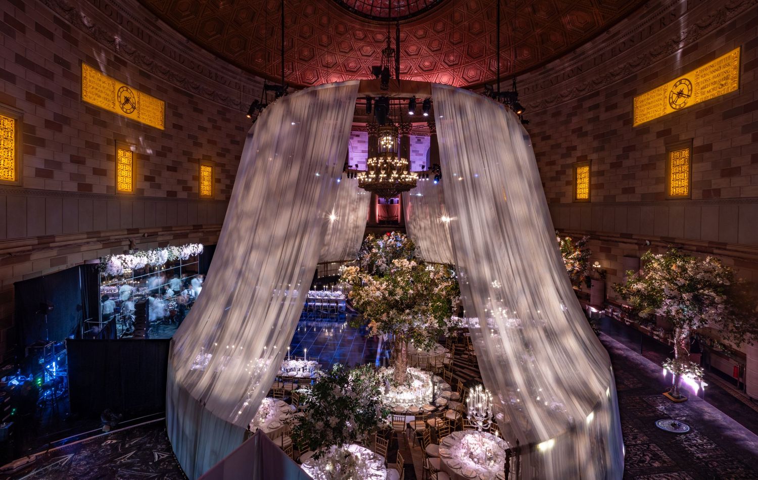Dramatic wedding reception setup with flowing white drapes, crystal chandelier and romantic mood lighting in brick venue.