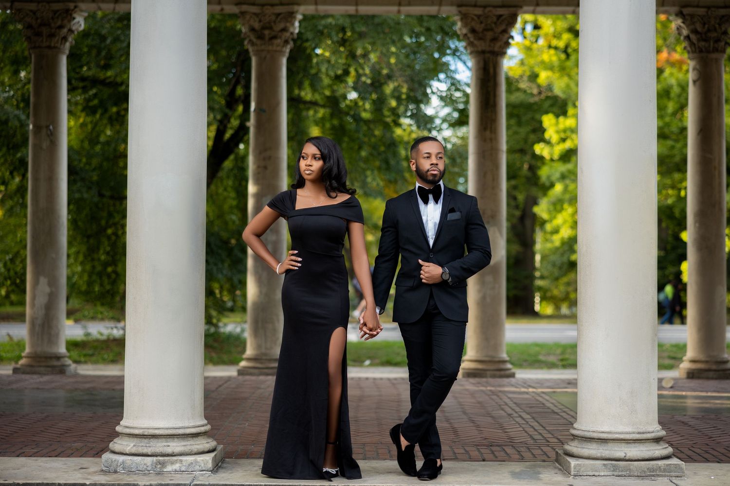 A couple in formal attire poses elegantly between white classical columns in an outdoor garden setting.